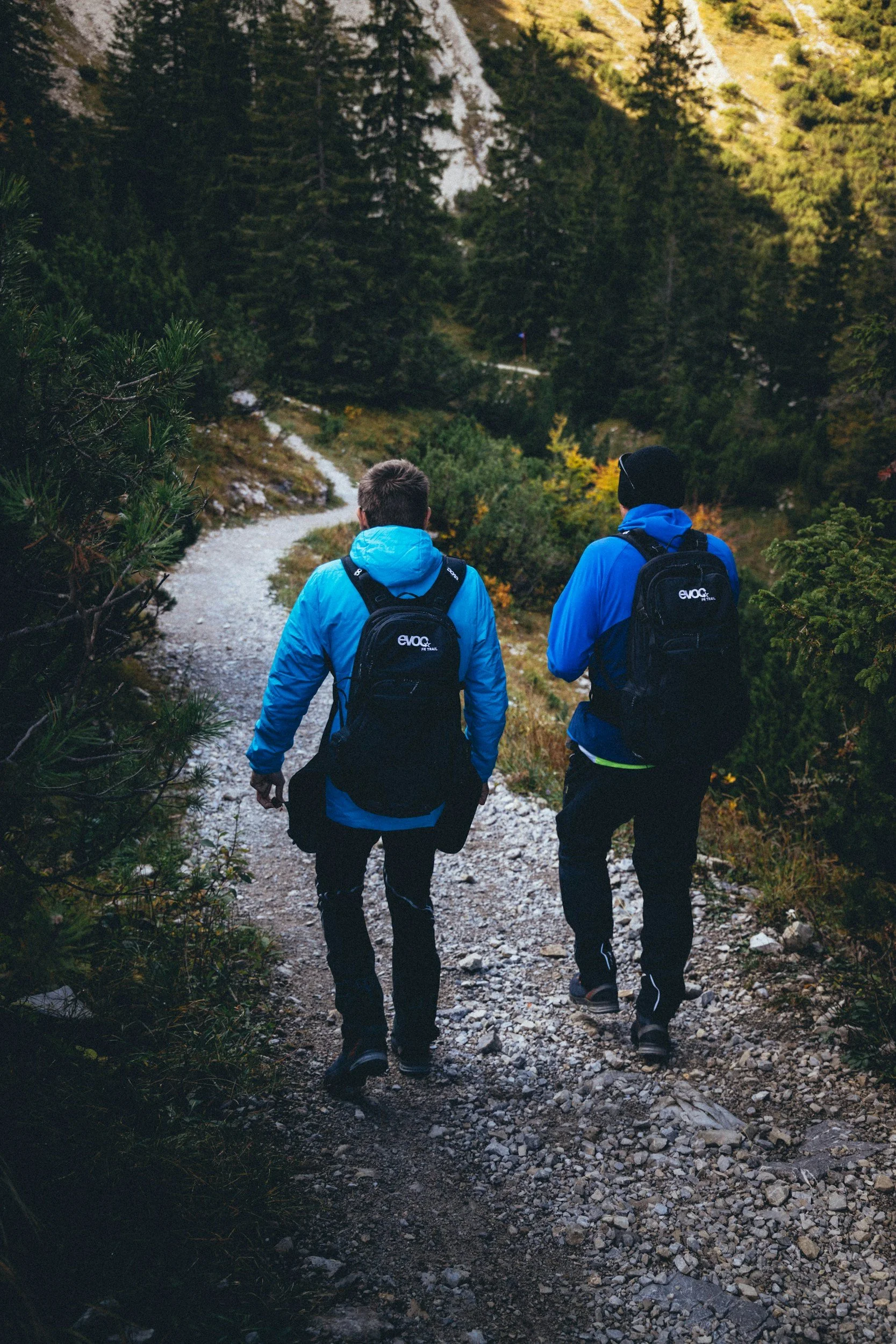 Two men in blue jackets and backpacks walk along a rocky path in a forest.
