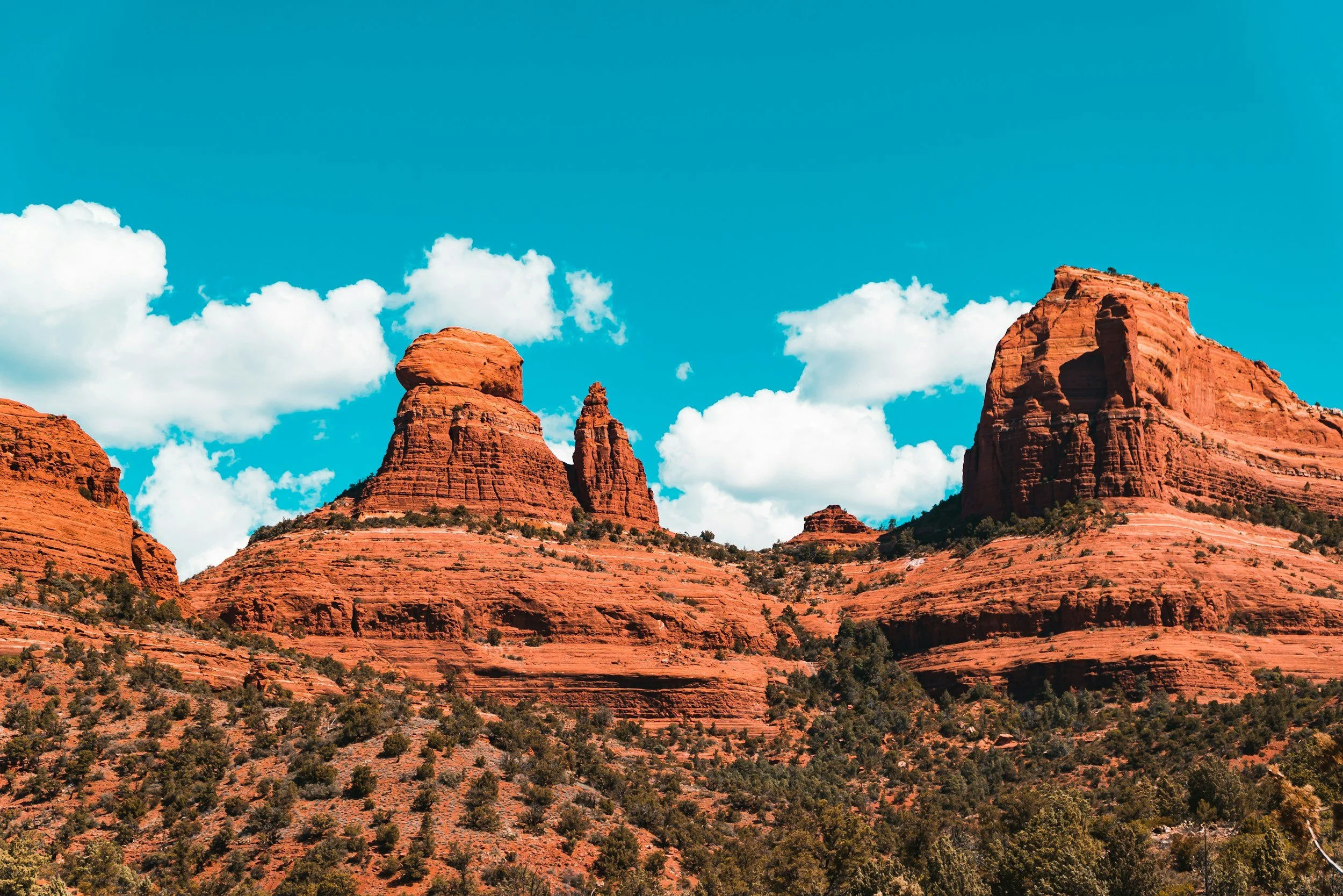 Red rocky outcrops and sparse vegetation of the Sedona, Arizona desert against a blue sky with puffy white clouds.