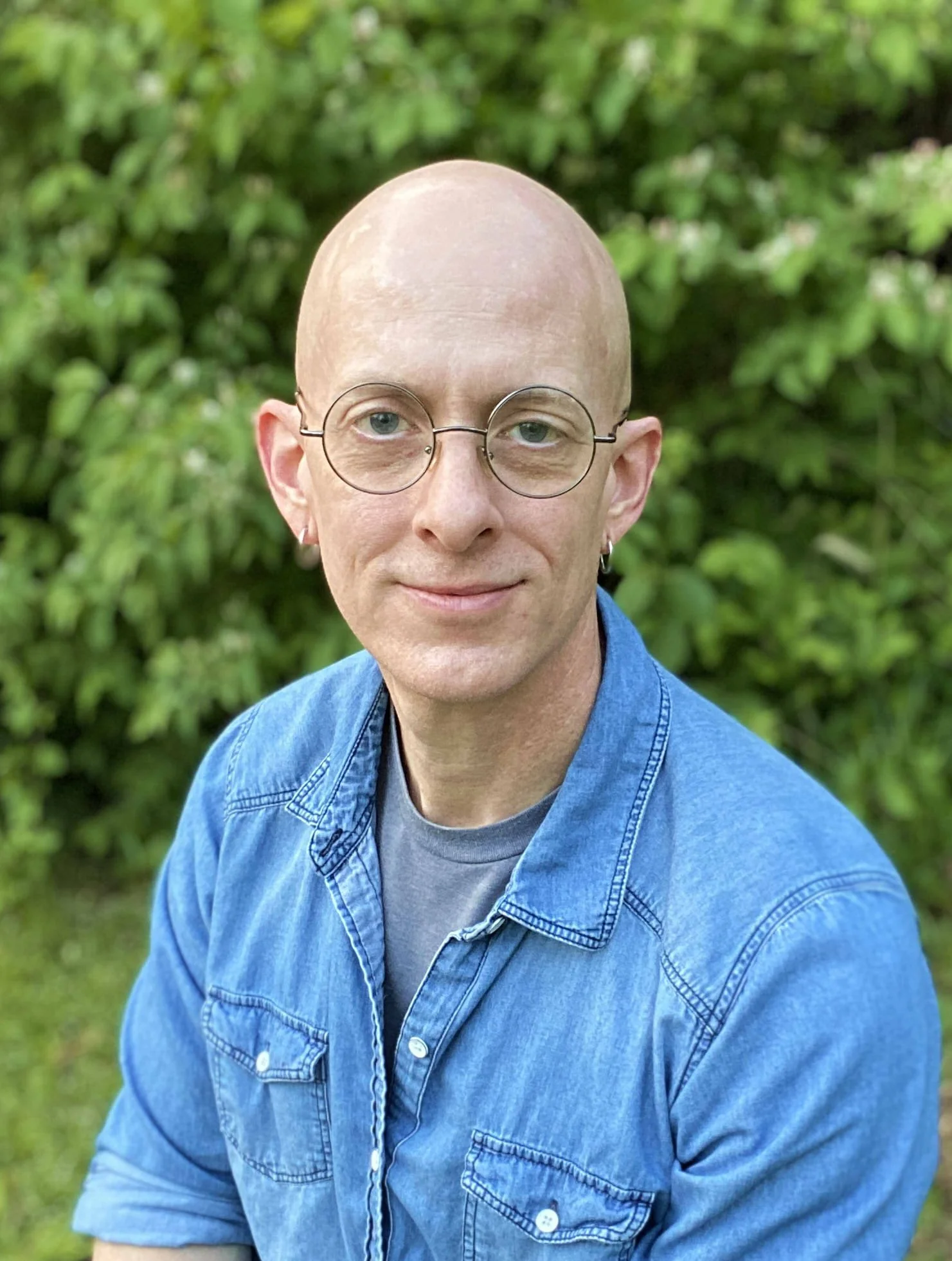 A therapist in Arkansas smiles softly. He wears round glasses, medium-gauge silver earrings, and a denim shirt.