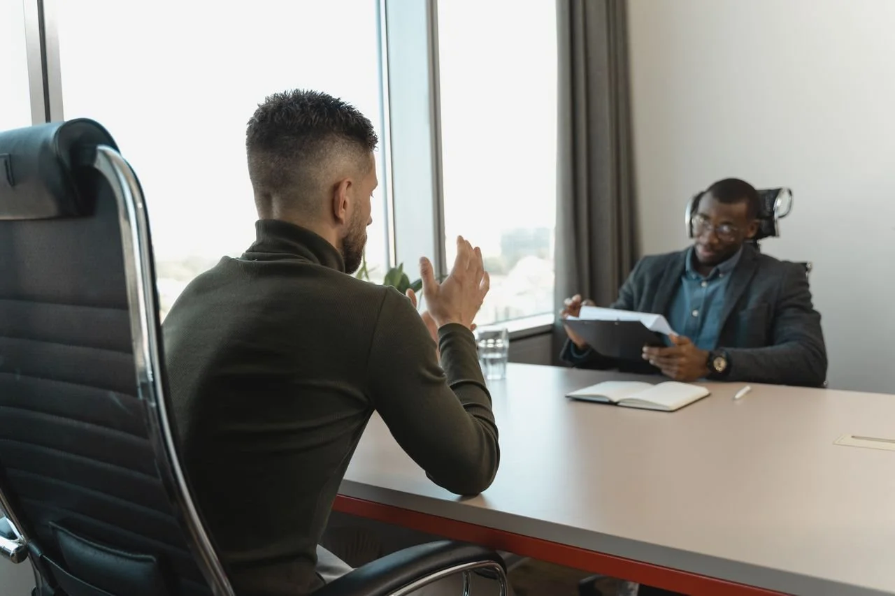 Man in turtleneck shirt gestures with his hands, talking to another man in a blazer on the other side of the desk. Photo by Tima Miroshnichenko courtesy of Pexels.