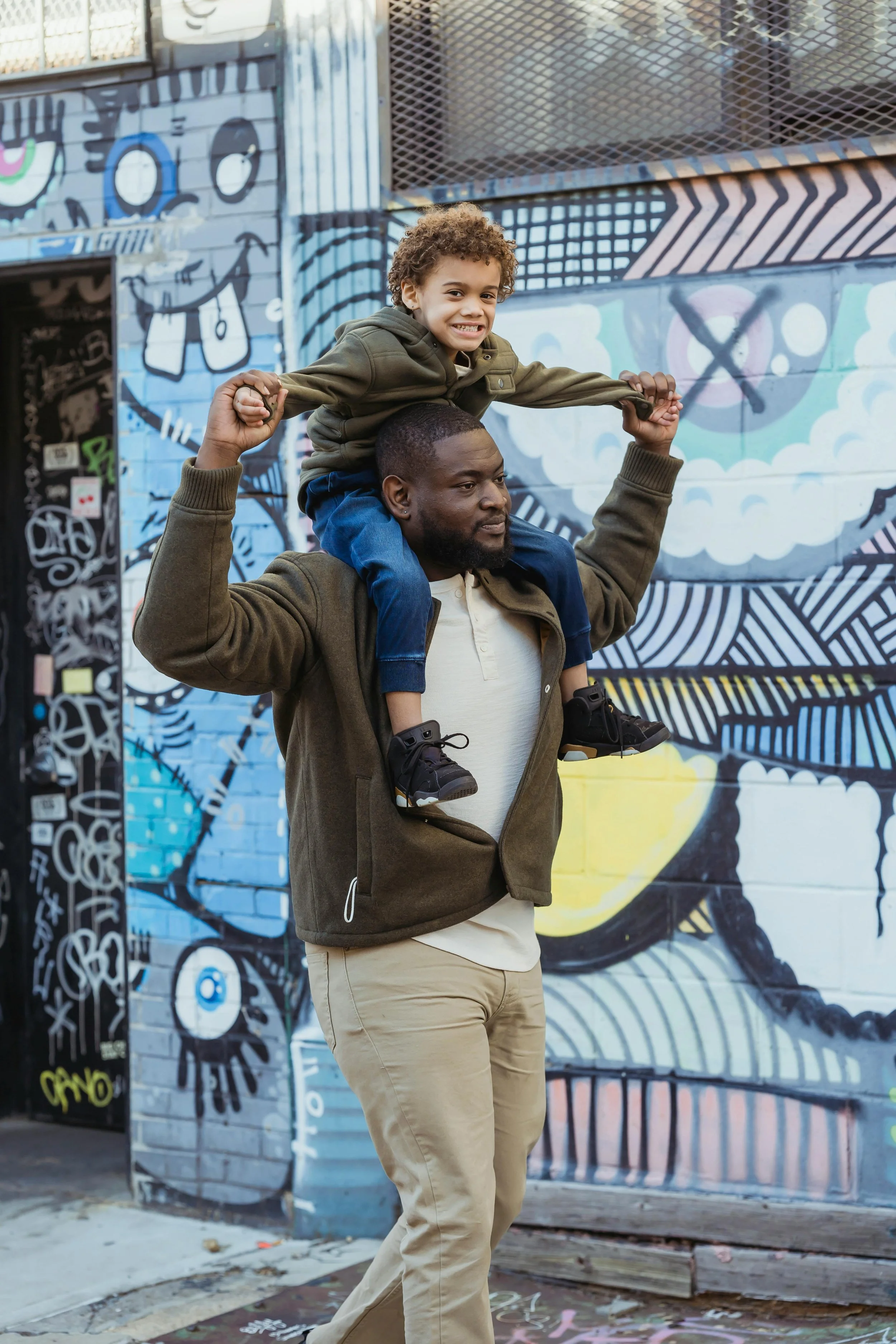 Man walks along urban street with mural carrying child on his shoulders. Photo by Kelra Burton.