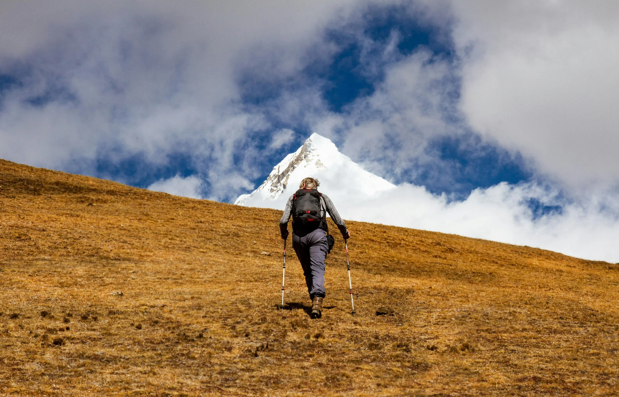 Man with backpack and trekking poles walks uphill with snowy mountain peak in distance. Photo by Tandln Bhutan.