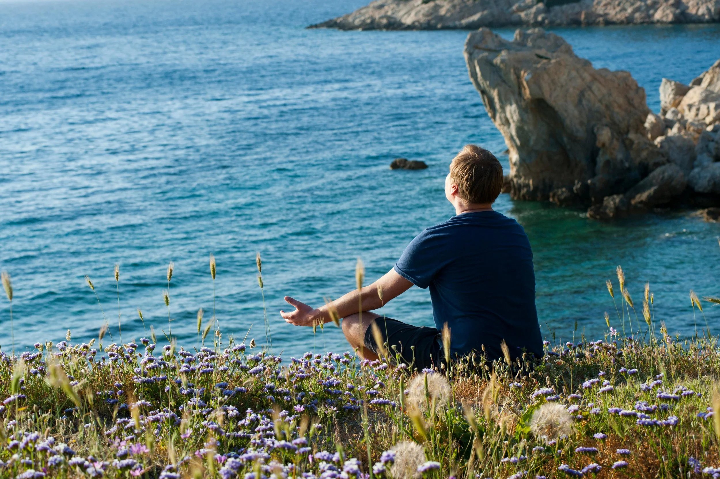 Man in blue shirt practices acceptance as he sits in meditation overlooking rocky ocean. Photo by Atlantic Ambience.