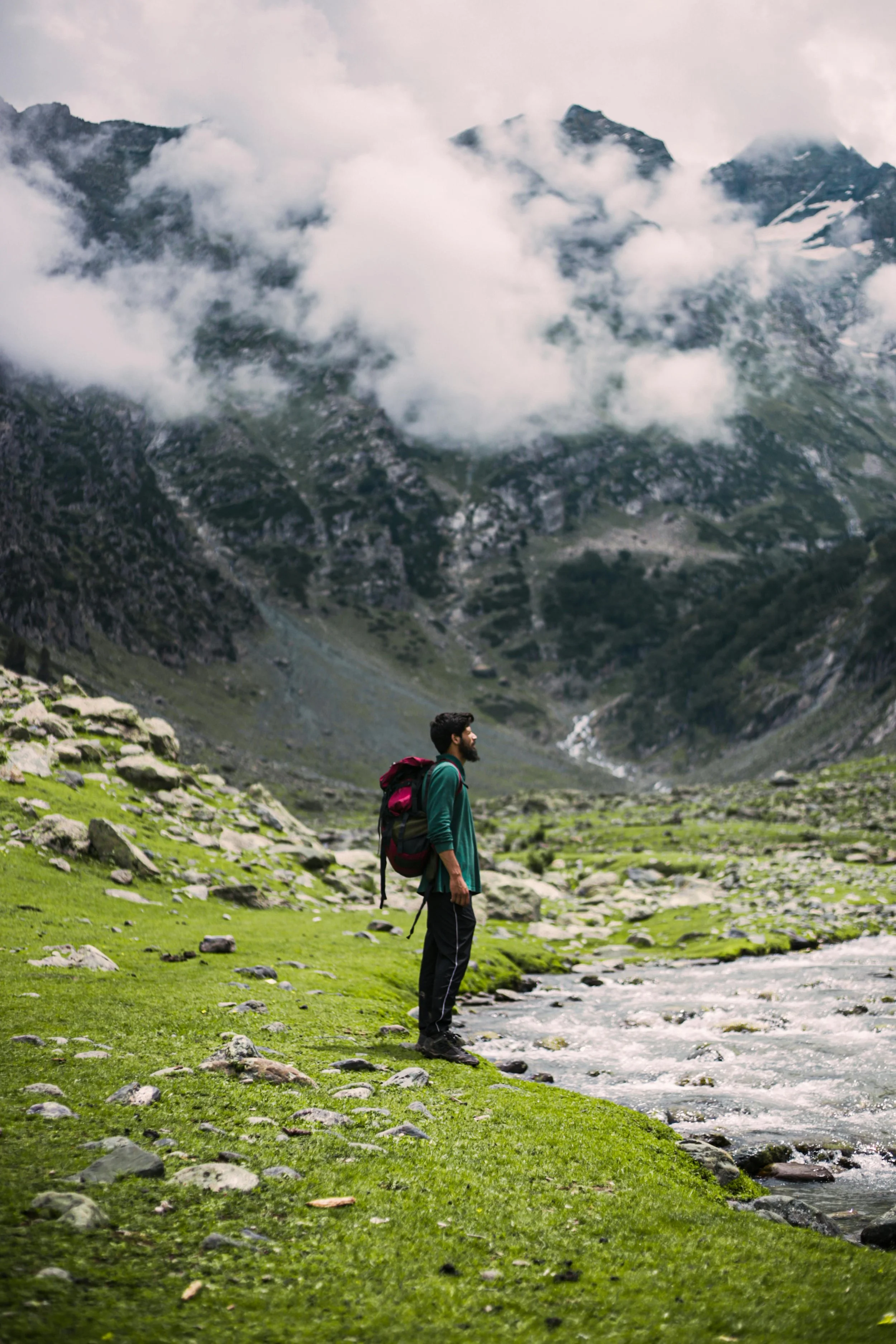 Man in green shirt with red backpack pauses at river edge. Tall mountains with clouds seen in background.
