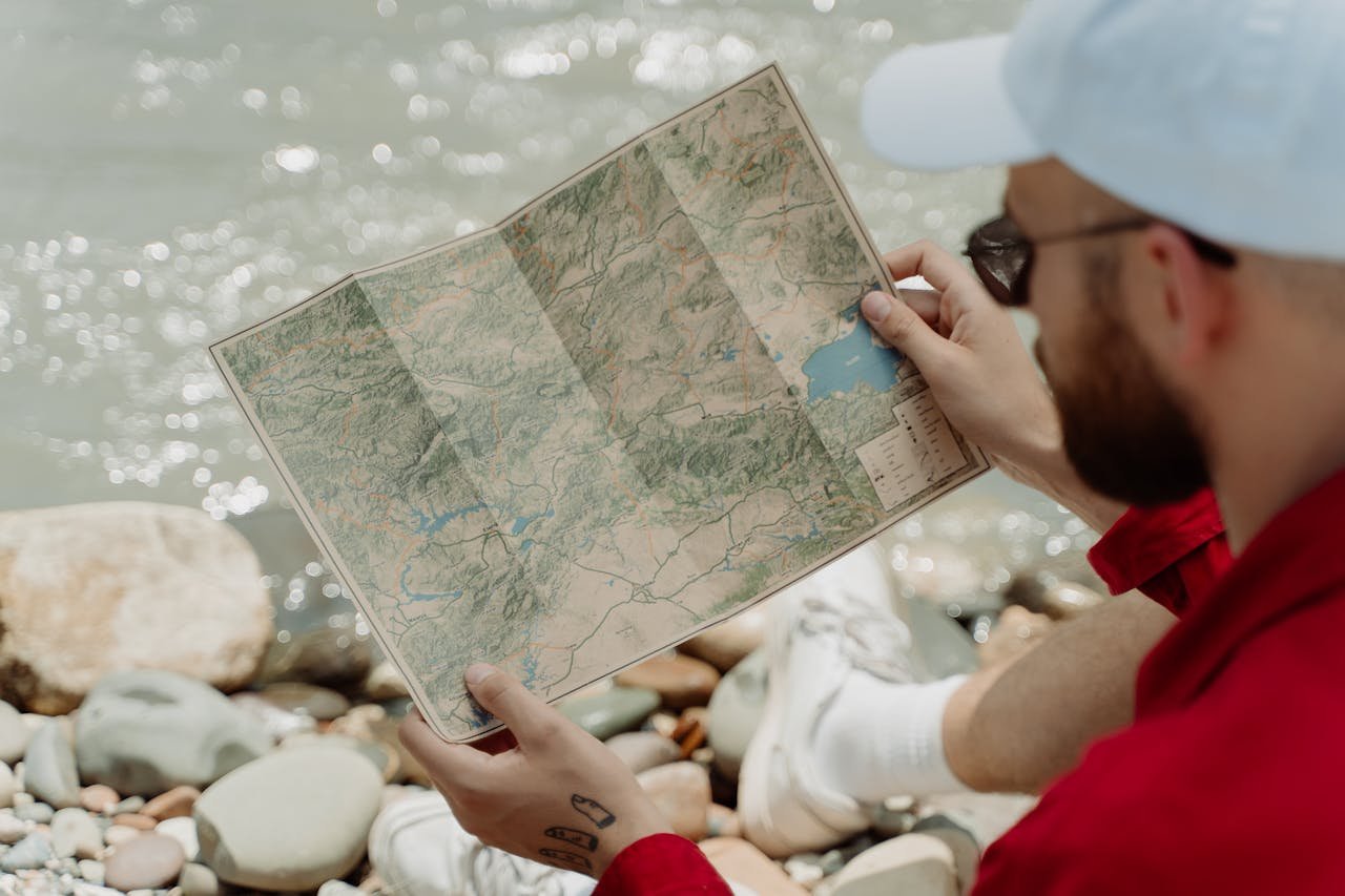 Bearded man in red jacket looks at map, sitting by stream. Photo by Cottonbro Sudio.