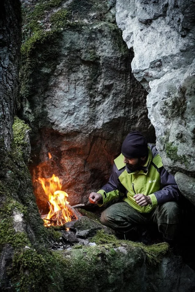 Bearded man in black and green jacket builds a fire tucked among rocks. Photo by Saleh Bakhshlyev.