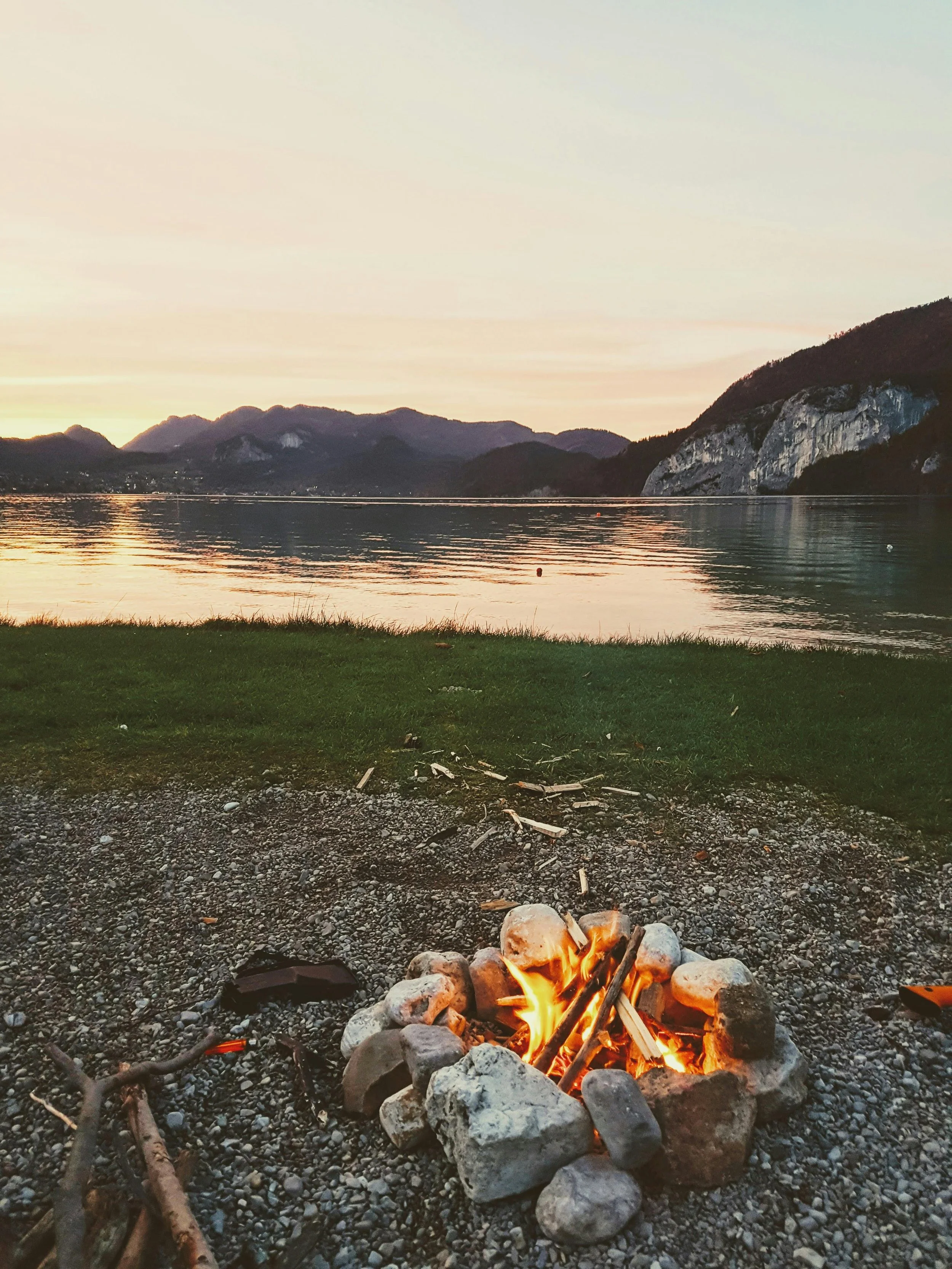 Small campfire in a circle of stones next to a lake with rocky hills in the distance