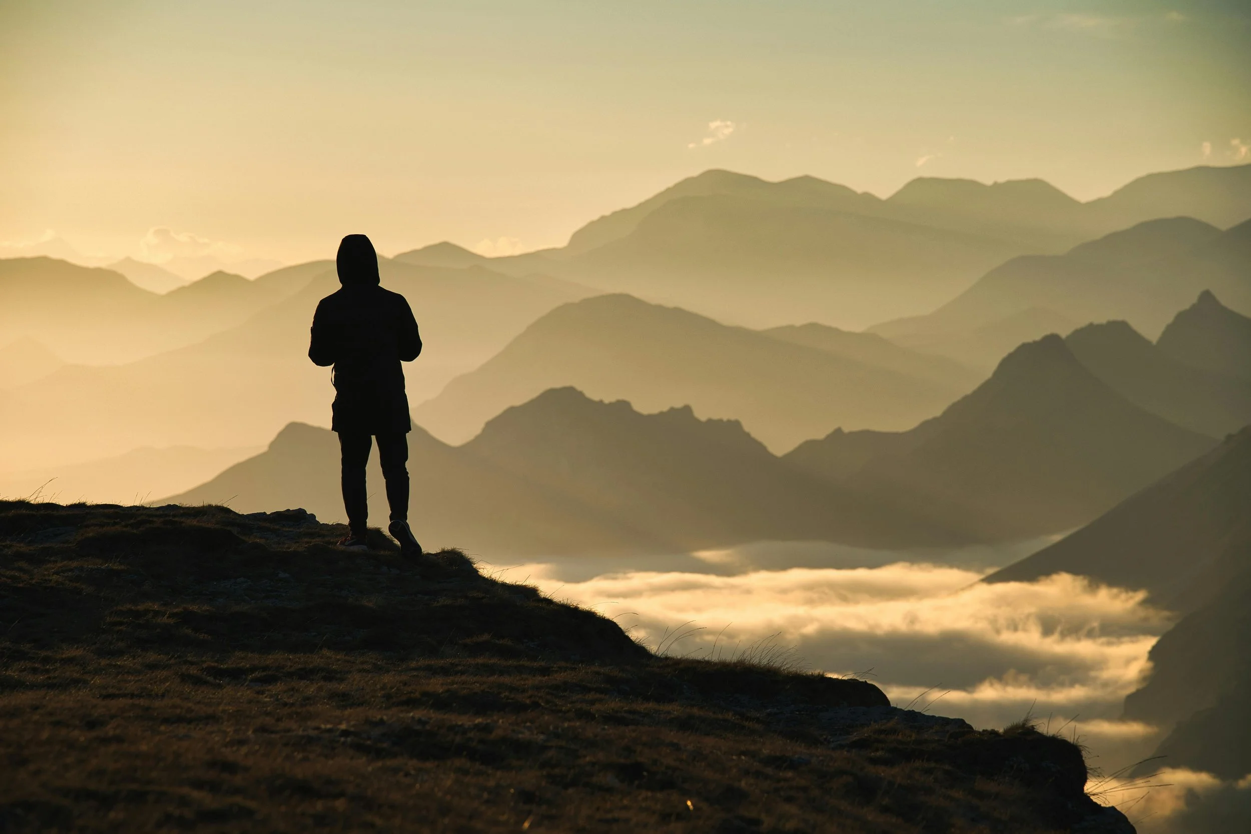 Silhouette of man on mountaintop overlooking cloudy mountains in the distance. Photo by Ivan Larin.