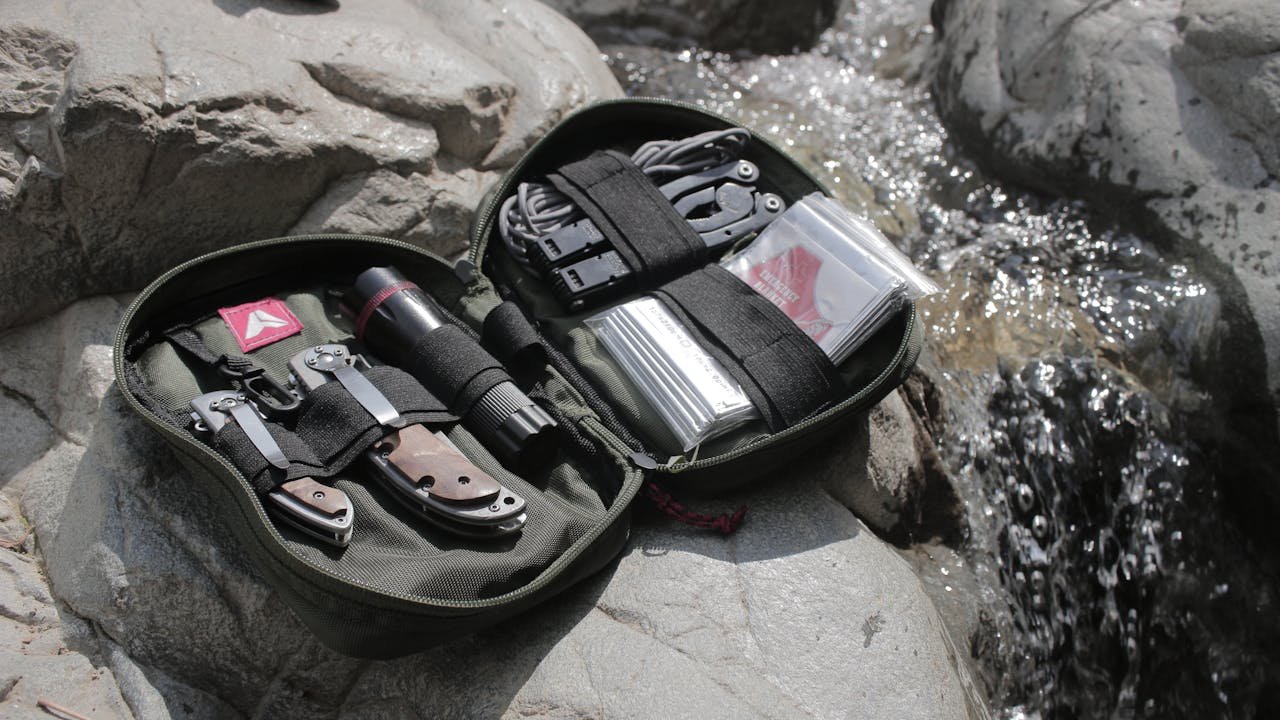 Olive green survival kit lays open near a small stream. Kit contains knives, flashlight, cord, pliers, and maps. Photo by Baihaki Hine.