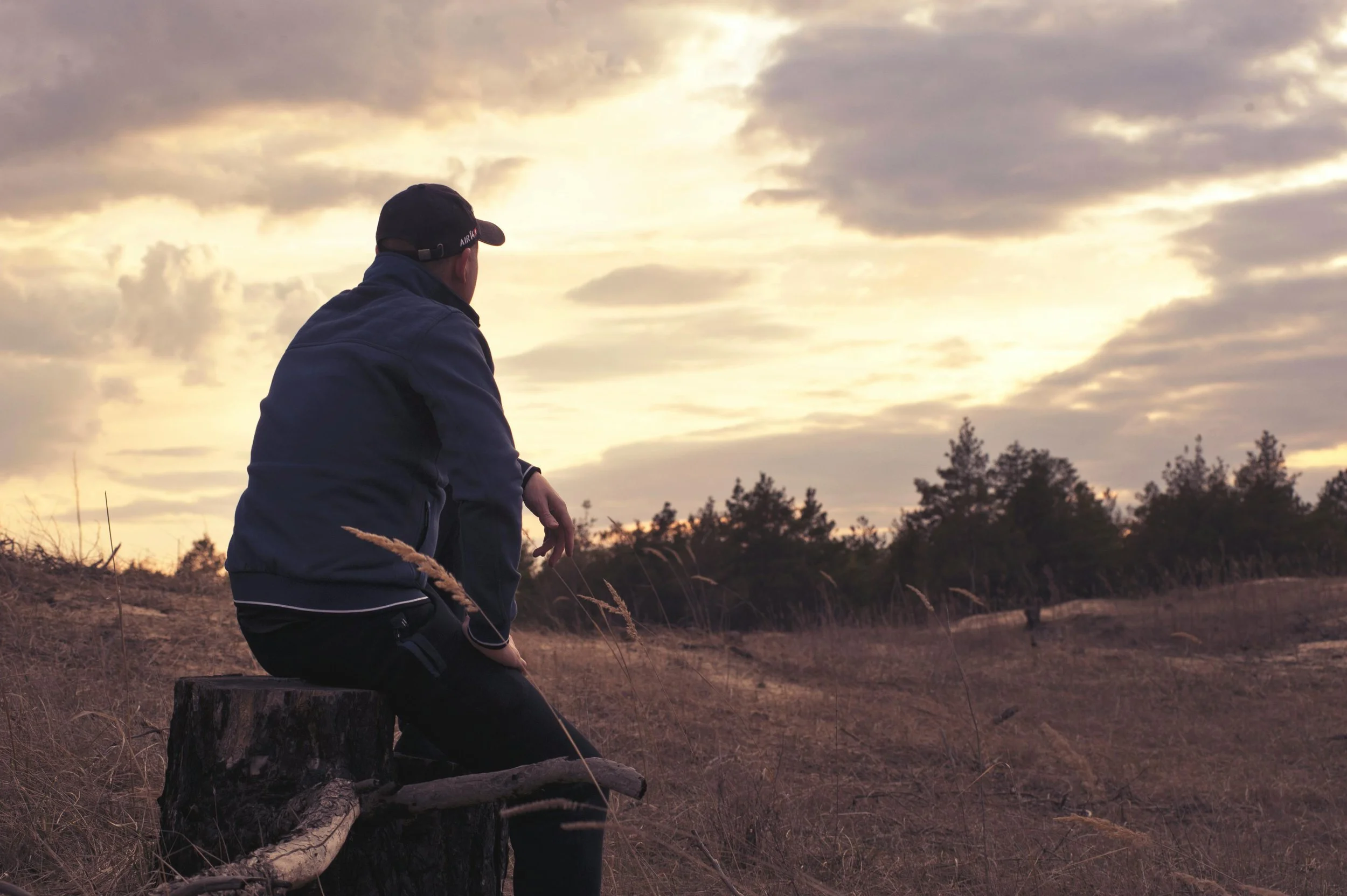 Man in ballcap and blue jacket sits alone on a stump in a field, looking at the sky
