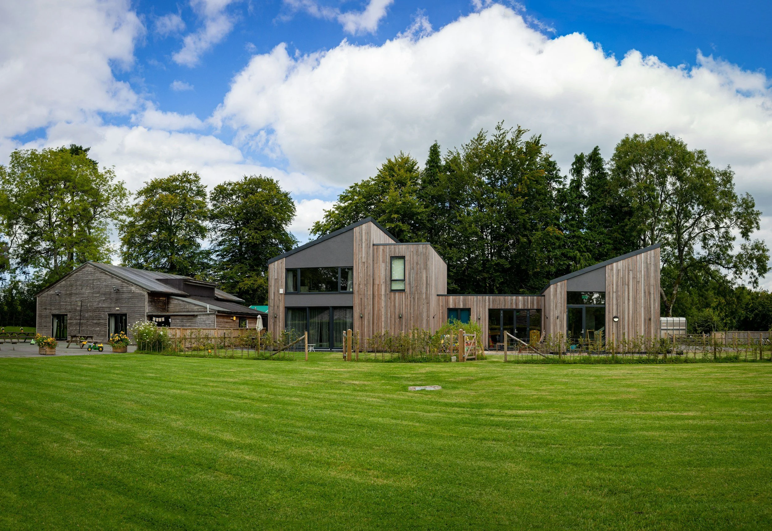 A modern house with a wooden exterior surrounded by a green lawn and trees under a partly cloudy sky.