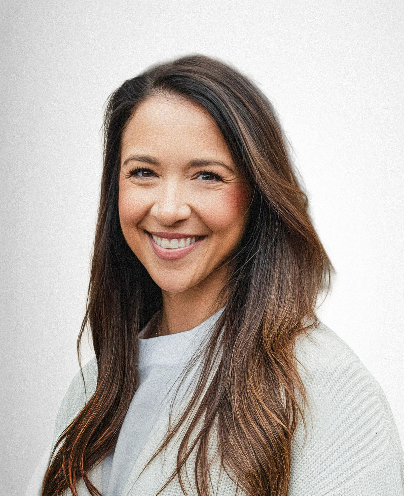 Close-up of a woman with long, wavy brown hair, smiling, wearing a white sweater, against a plain light background.