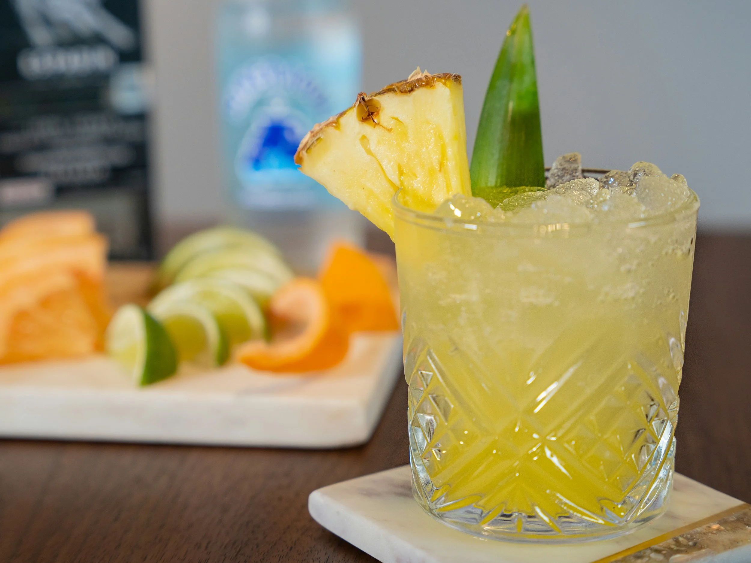 A yellow cocktail garnished with a pineapple slice and a green leaf, served in a decorative glass with crushed ice.