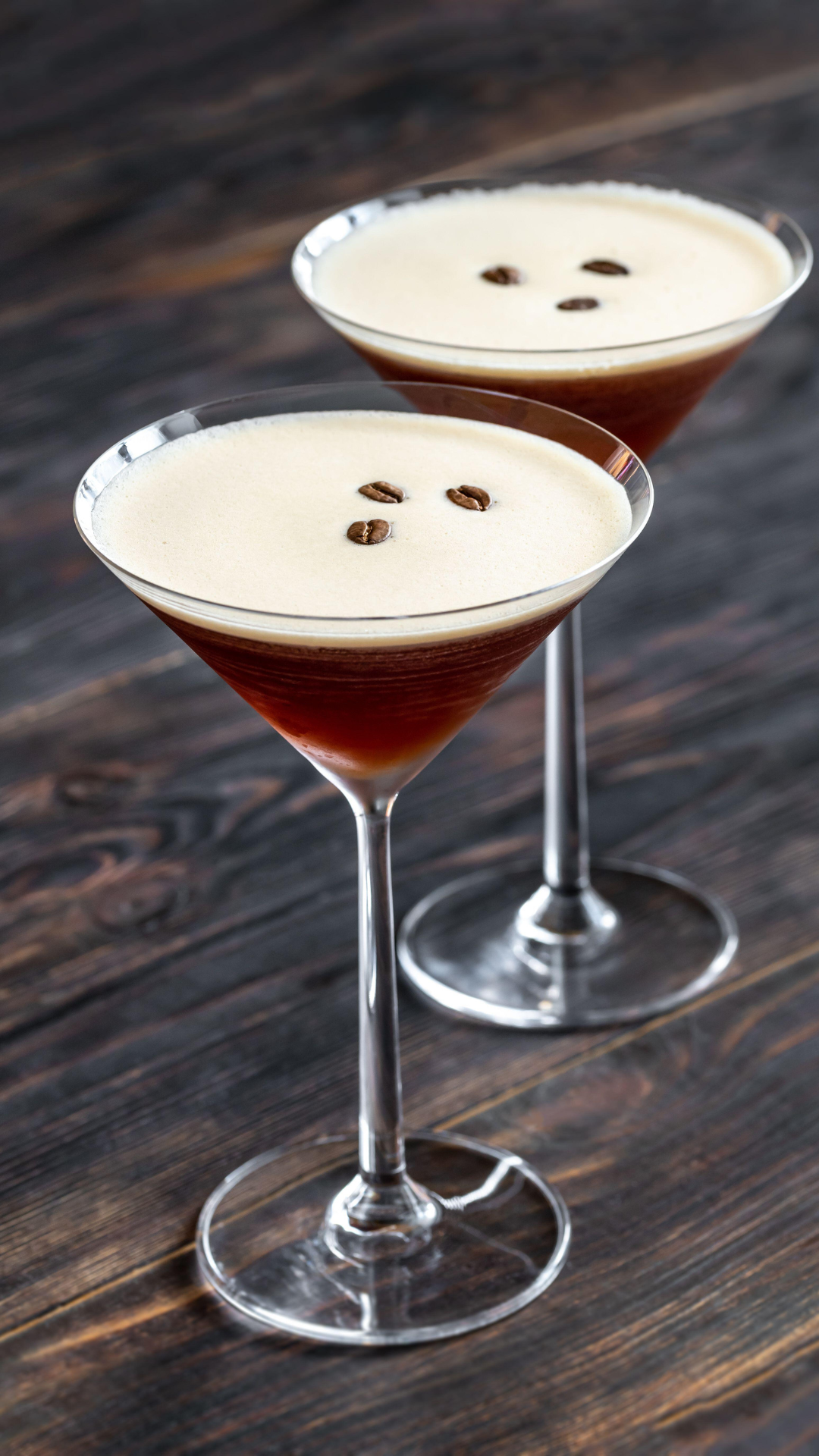 Two martini glasses filled with coffee cocktails topped with cream and garnished with coffee beans, set on a dark wooden surface.
