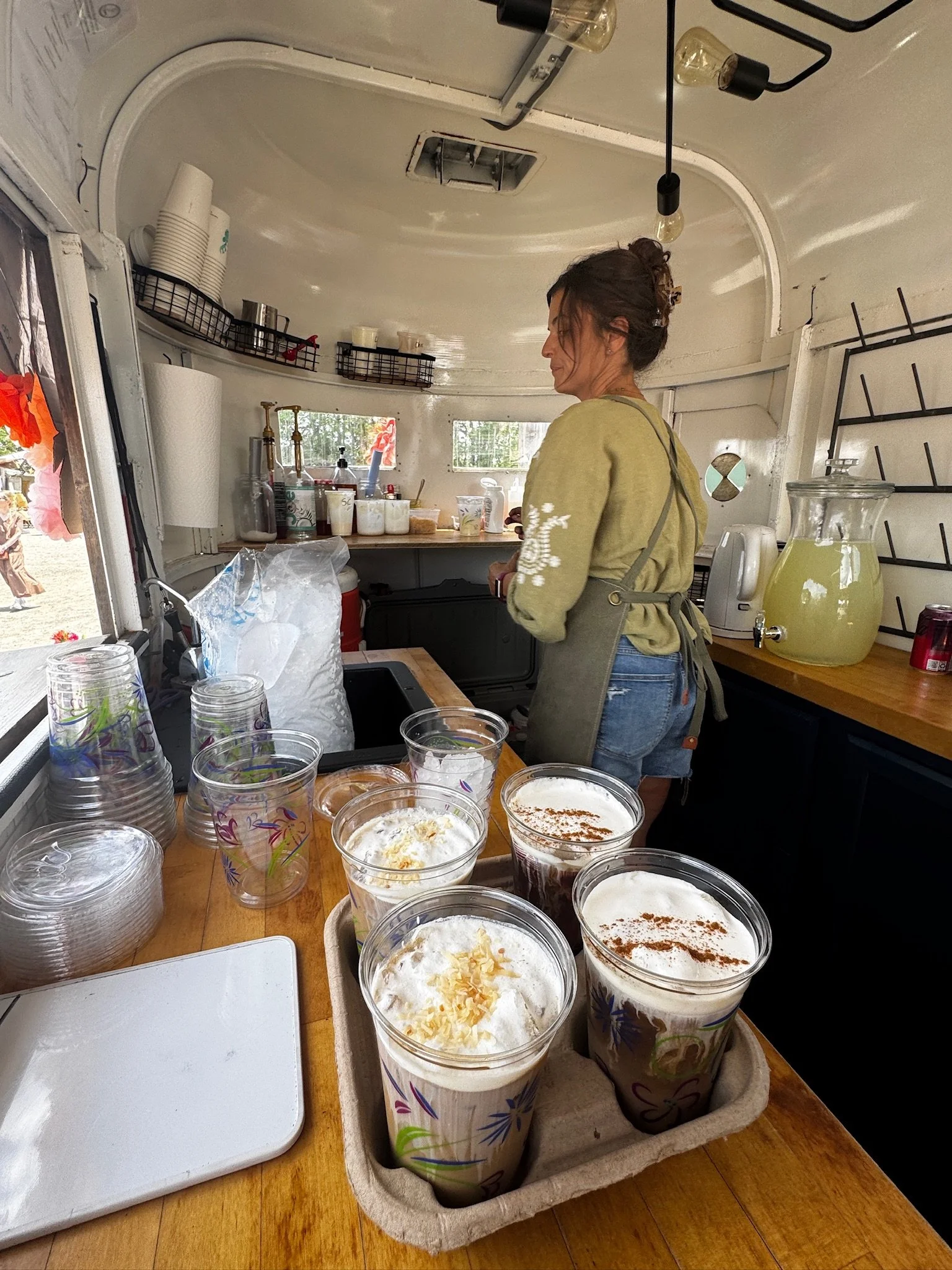 A woman in a green sweatshirt and apron preparing drinks inside a small food truck or stand. Multiple cups of frozen beverages with whipped cream and toppings are on a tray in the foreground. The interior has shelves with cups, bottles, and supplies, and a large pitcher of lemonade on the counter.