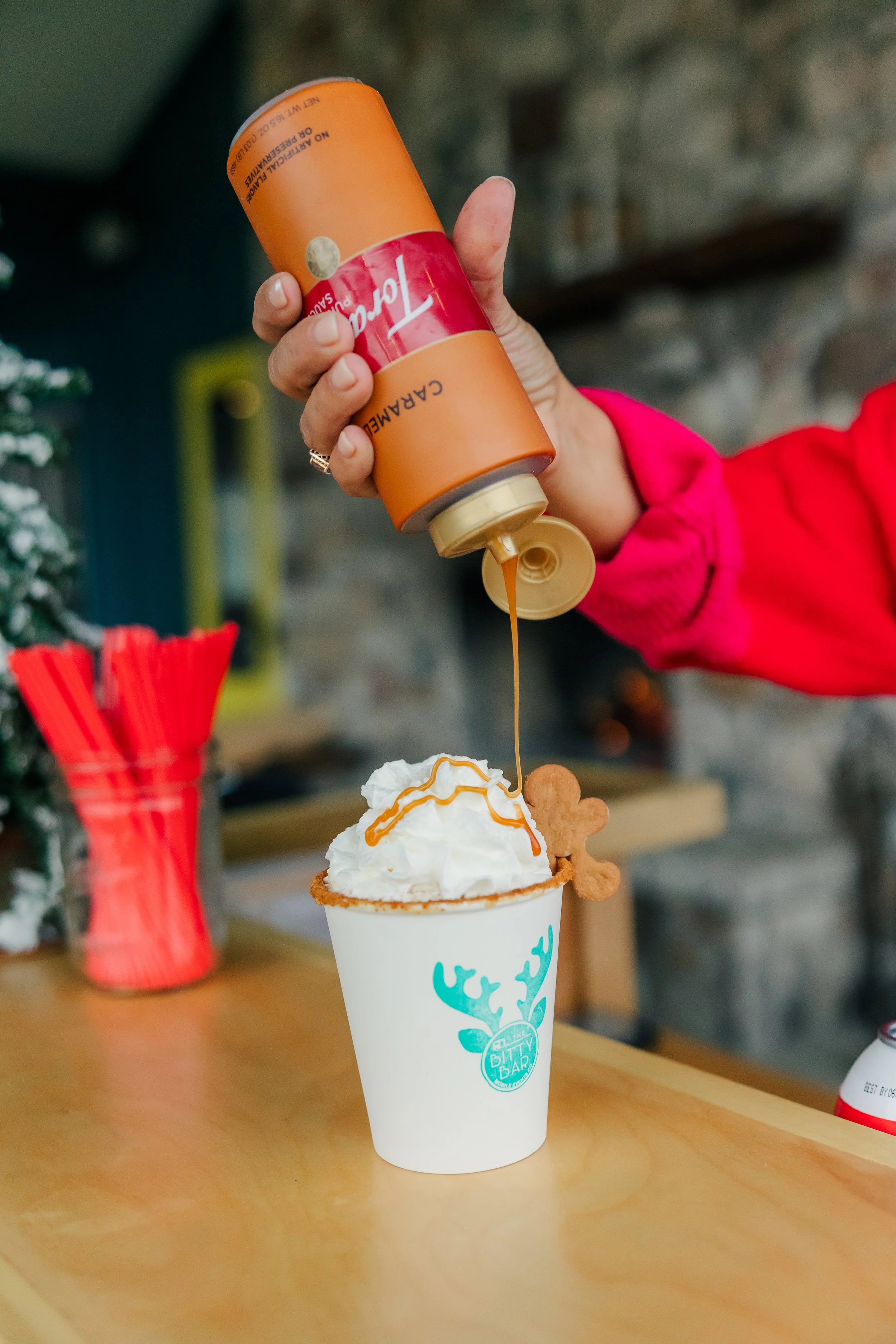 The Bitty Bar bartender holding caramel sauce over a cup of whipped cream-topped Hot Cocoa with gingerbread cookies on the rim. In the background, there are red straws in a container and a blurred indoor setting.