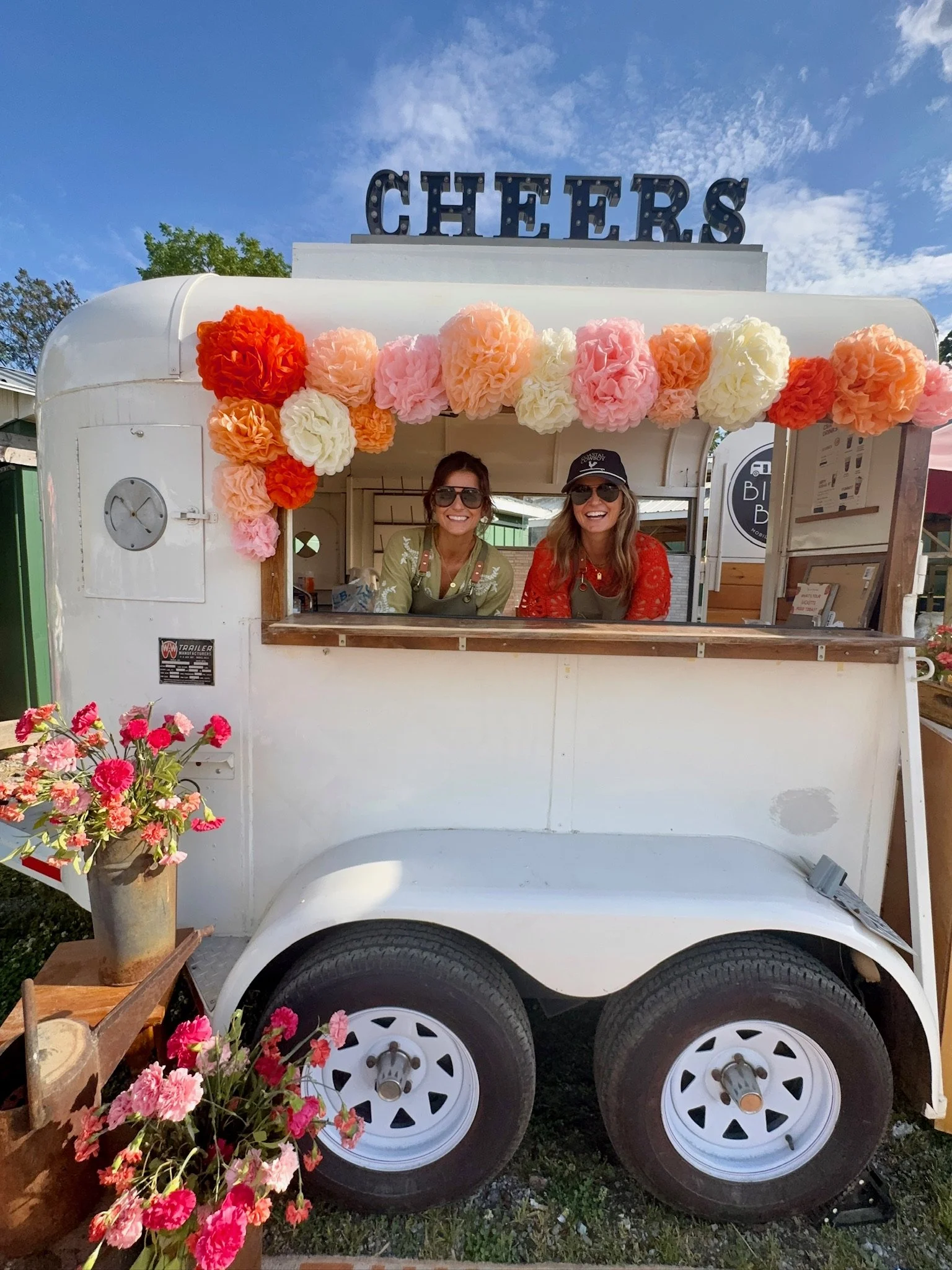 A vintage white food trailer decorated with pink, peach, and white paper pom-poms and flowers. Two smiling women are standing inside the trailer, wearing sunglasses. There are flowers in a pot in the foreground and a sign that says "CHEERS" is mounted on top of the trailer. The sky is blue with some clouds.