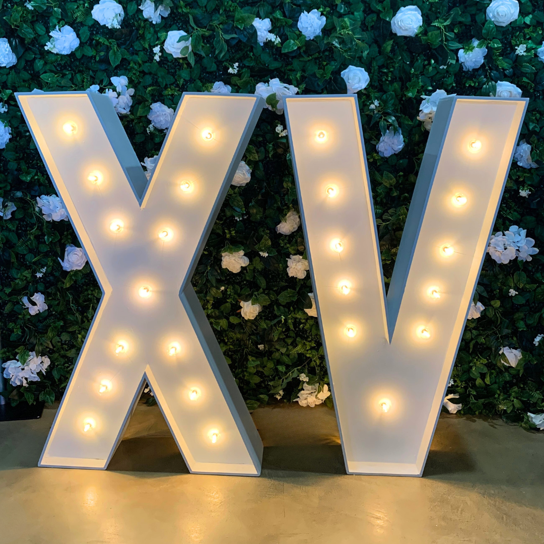 Large illuminated letterings spelling 'X V' against a background of white flowers and green leaves.