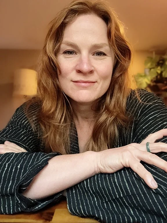 Close-up of a woman with long, wavy red hair, smiling softly, resting her arms on a table, wearing a dark pinstripe shirt, in a warmly lit indoor setting.