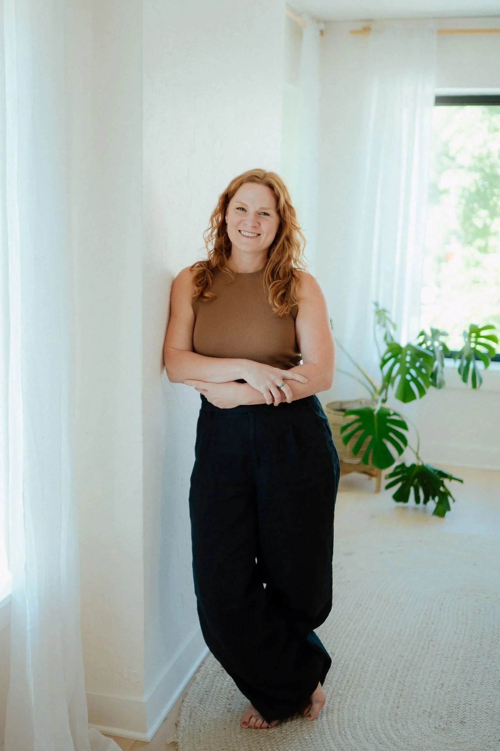 A woman with red hair standing in a cozy, naturally lit room. She is smiling, wearing a brown sleeveless top and black pants, with her arms crossed. The background features a large green leafy plant, white curtains, and a window.