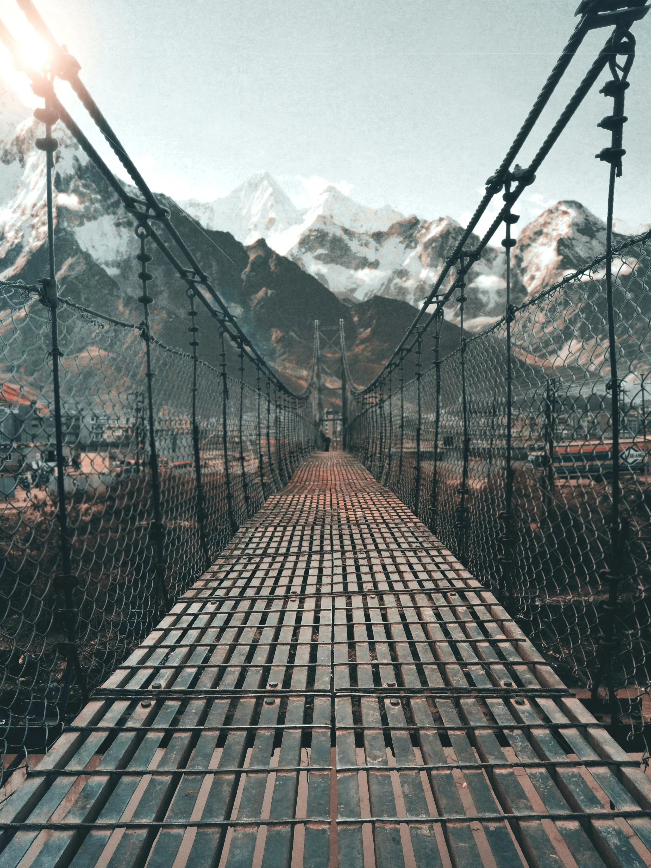 A metal suspension bridge with wooden planks stretches towards snow-capped mountains in the distance under a clear sky.