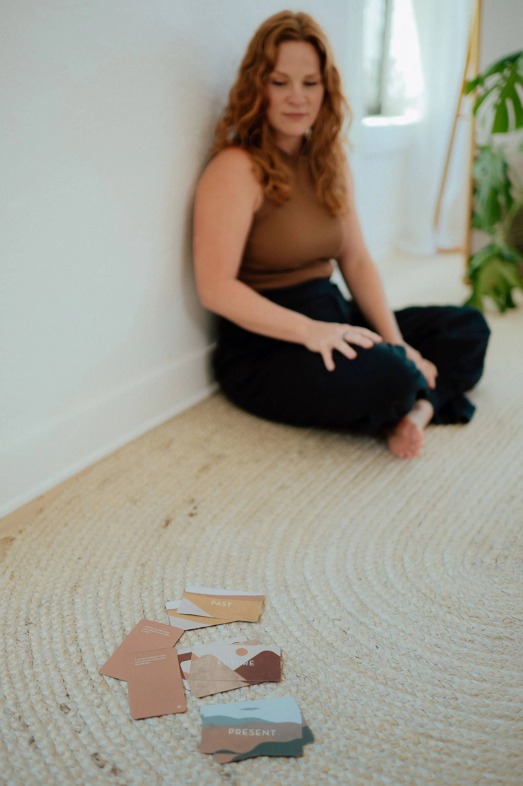 A woman with red hair sitting on the floor against a white wall, with scattered cards featuring words like 'PAST' and 'PRESENT' in front of her.