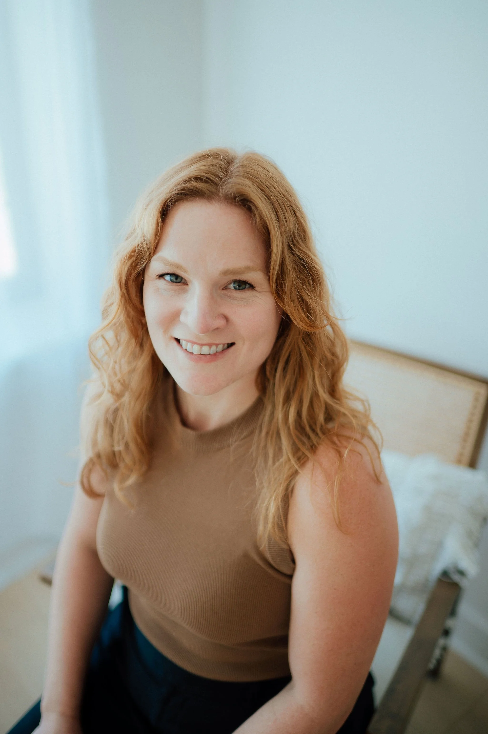 A smiling woman with curly red hair sitting on a bed in a softly lit room, wearing a beige sleeveless top and dark pants.