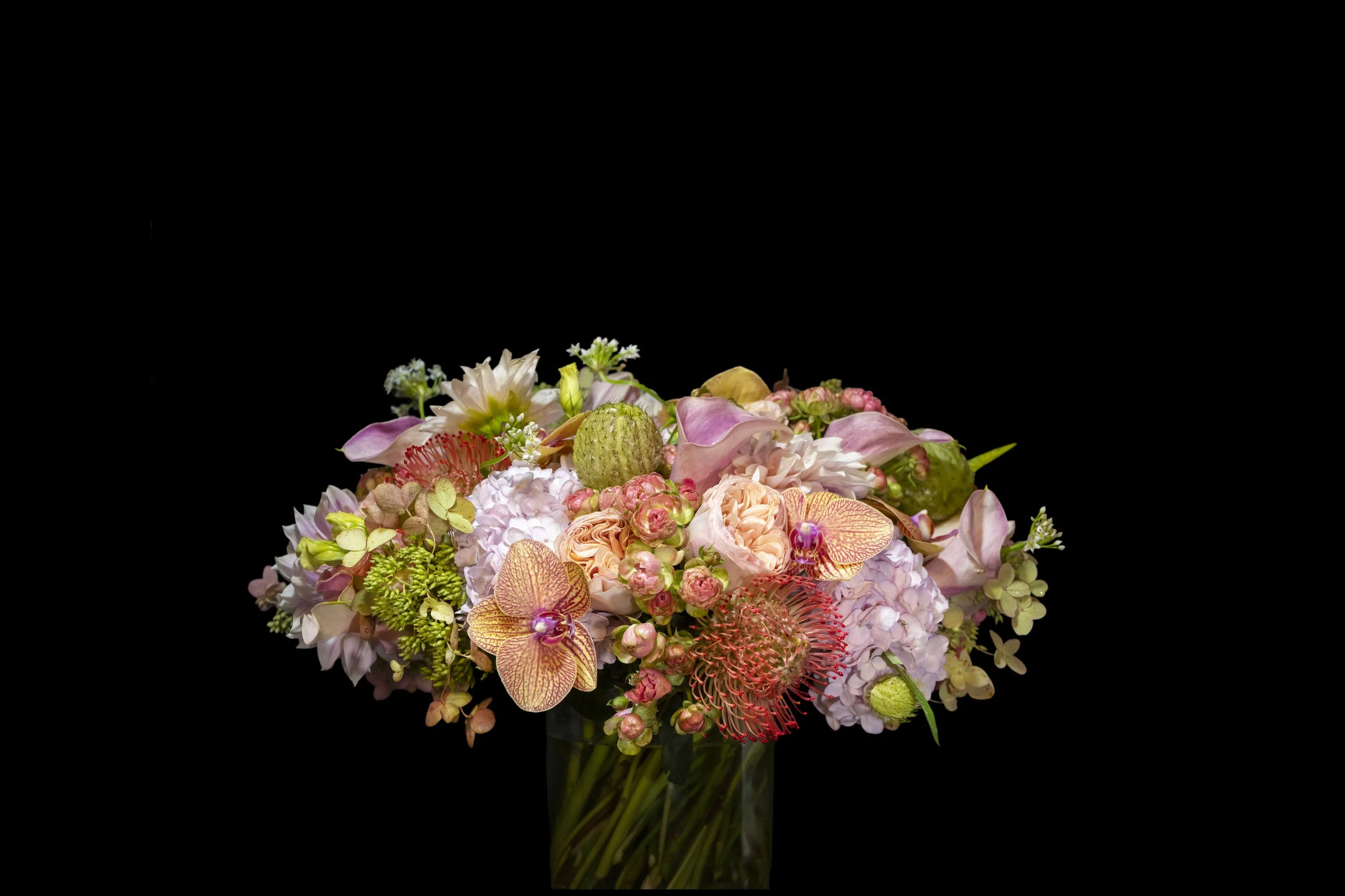A bouquet of various flowers including pink roses, orange orchids, and yellow-green accents, arranged against a dark background.