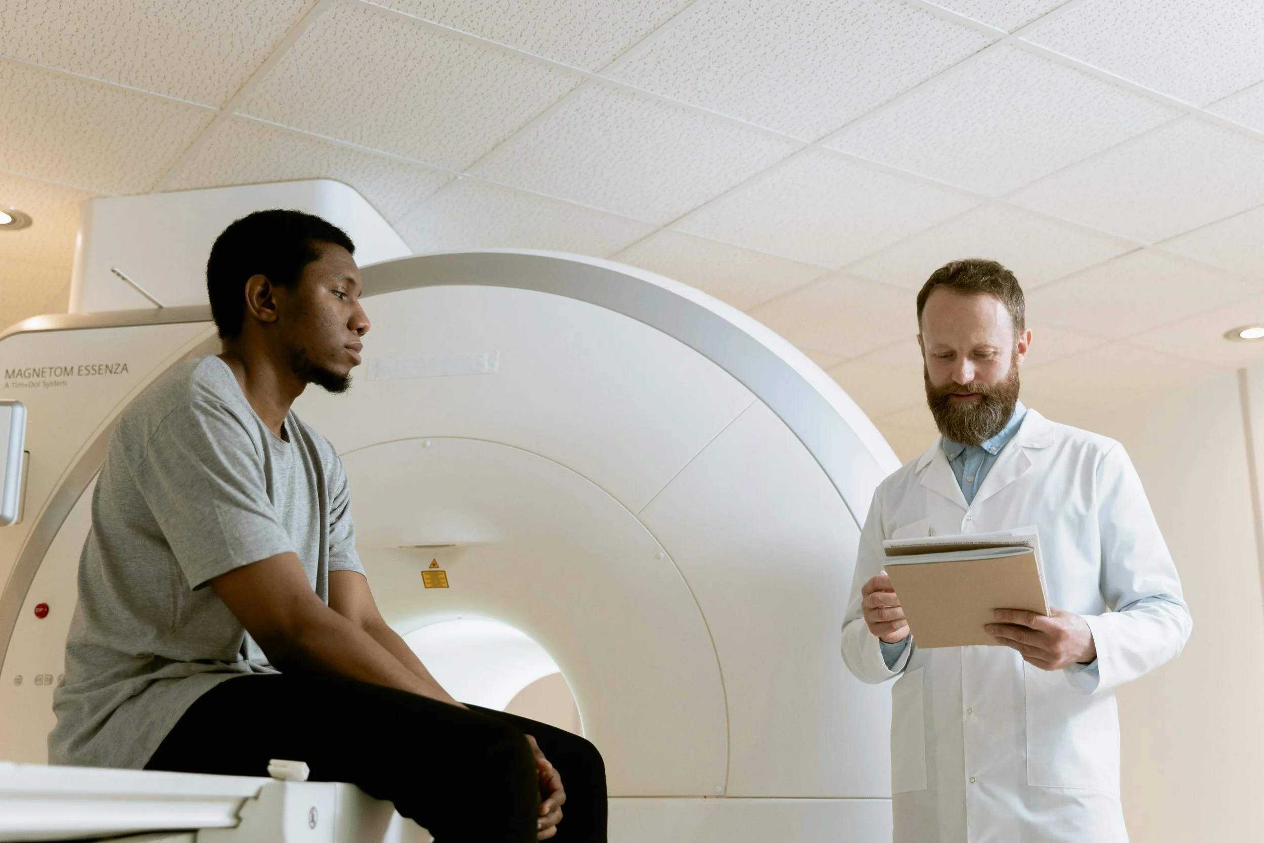 A man sitting on a hospital bed with an MRI machine behind him, talking to a doctor who is holding a notepad and looking at him, in a hospital room.