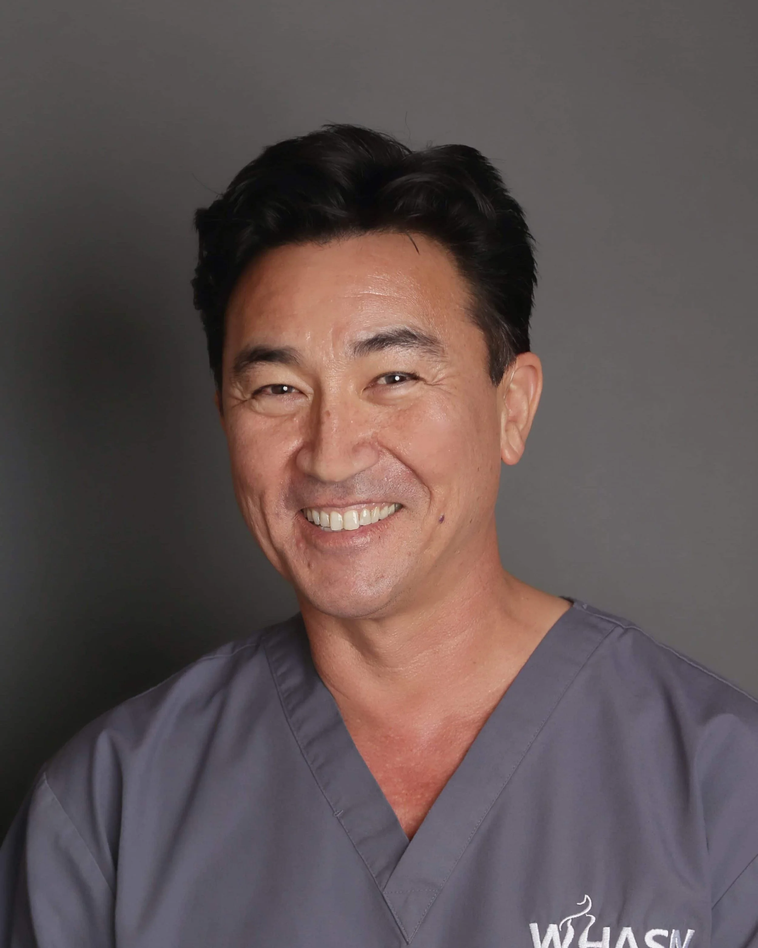 A man wearing gray medical scrubs with a WHASN logo, smiling against a gray background.