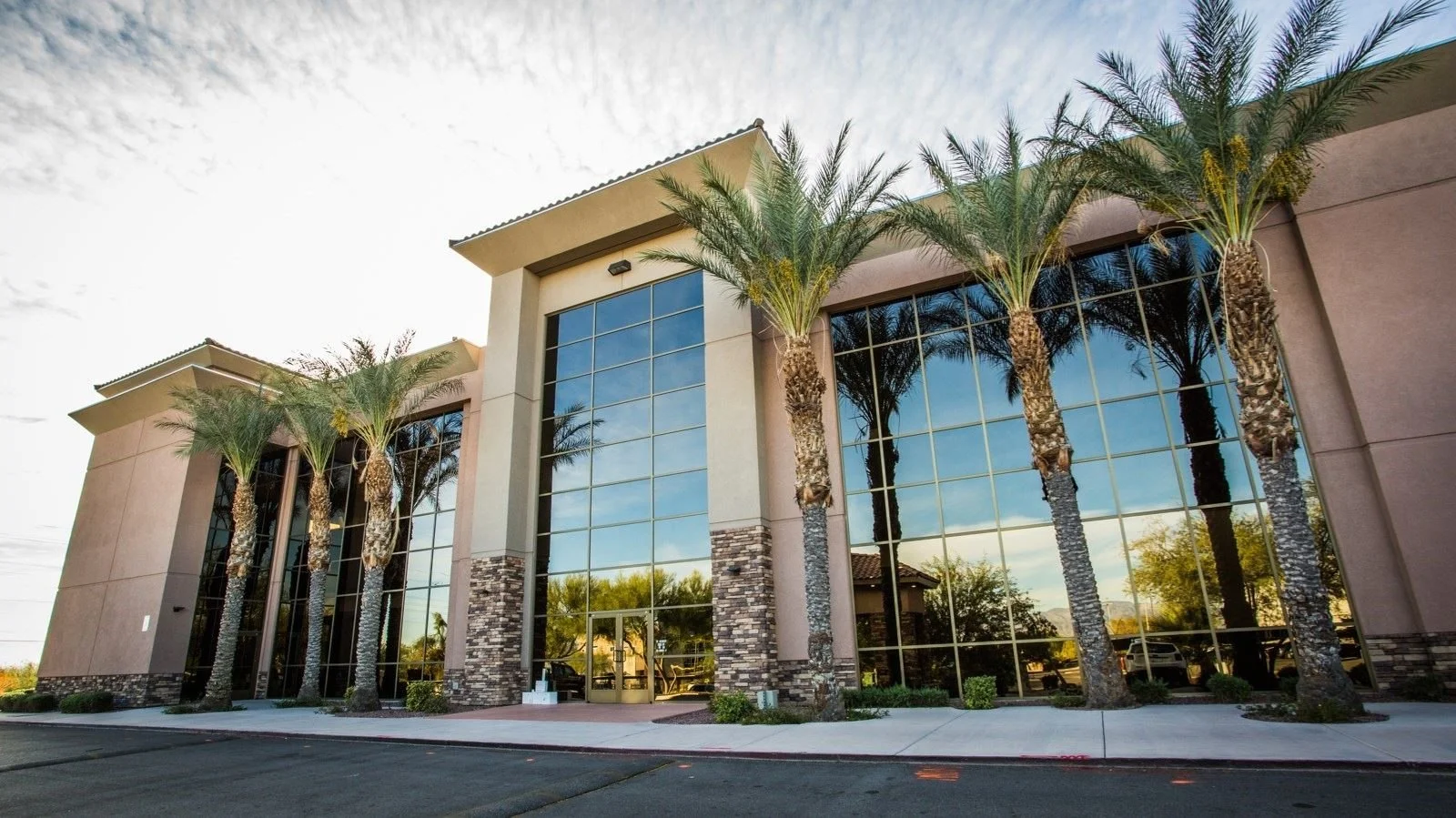 Modern commercial building with large glass windows, beige walls, and four tall palm trees in front.
