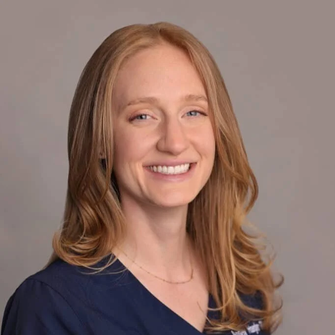 A young woman with long, wavy red hair smiling at the camera, wearing a dark navy scrubs top and a delicate gold necklace against a neutral grey background.