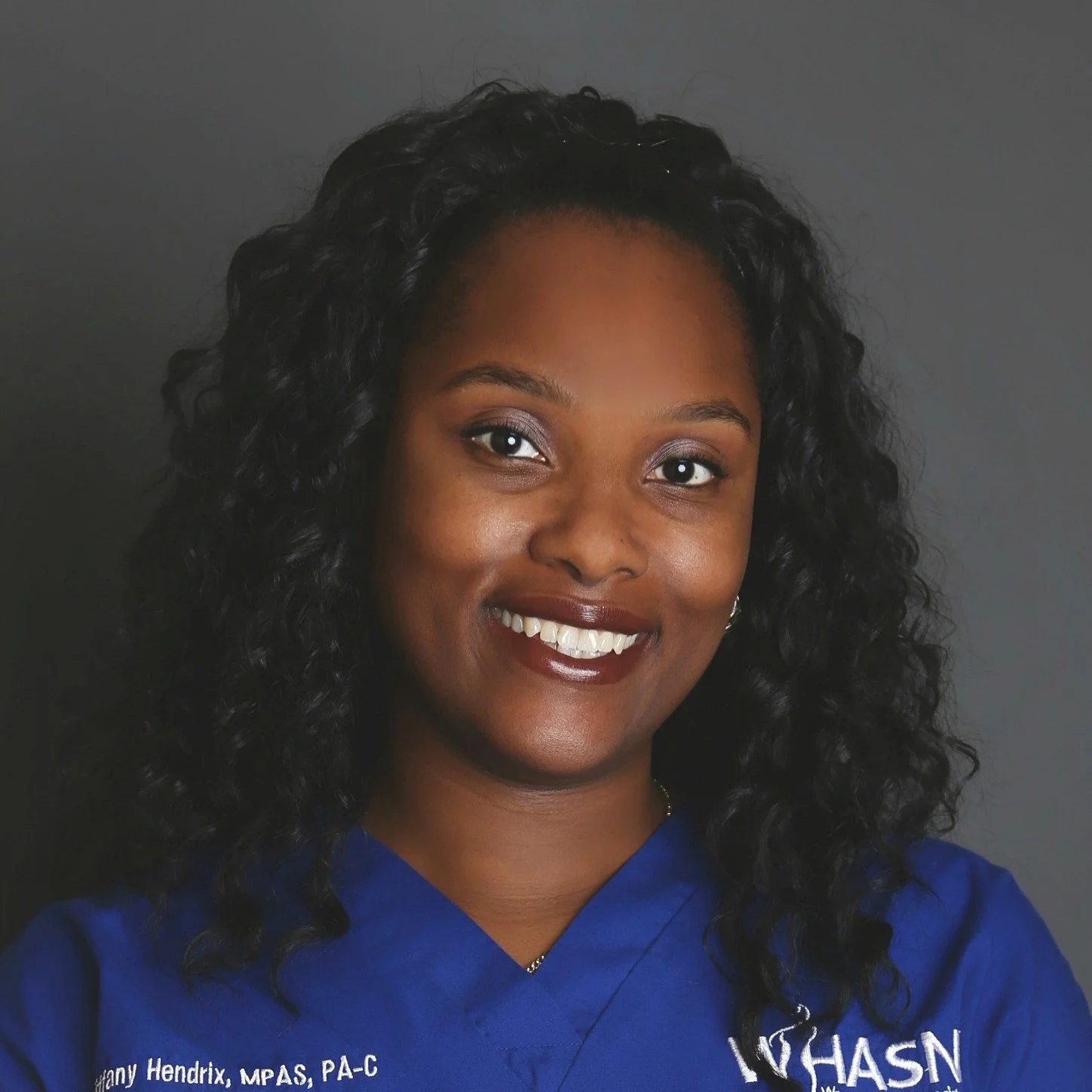 A woman with dark curly hair and a bright smile, wearing a blue medical uniform with embroidered text, standing against a gray background.