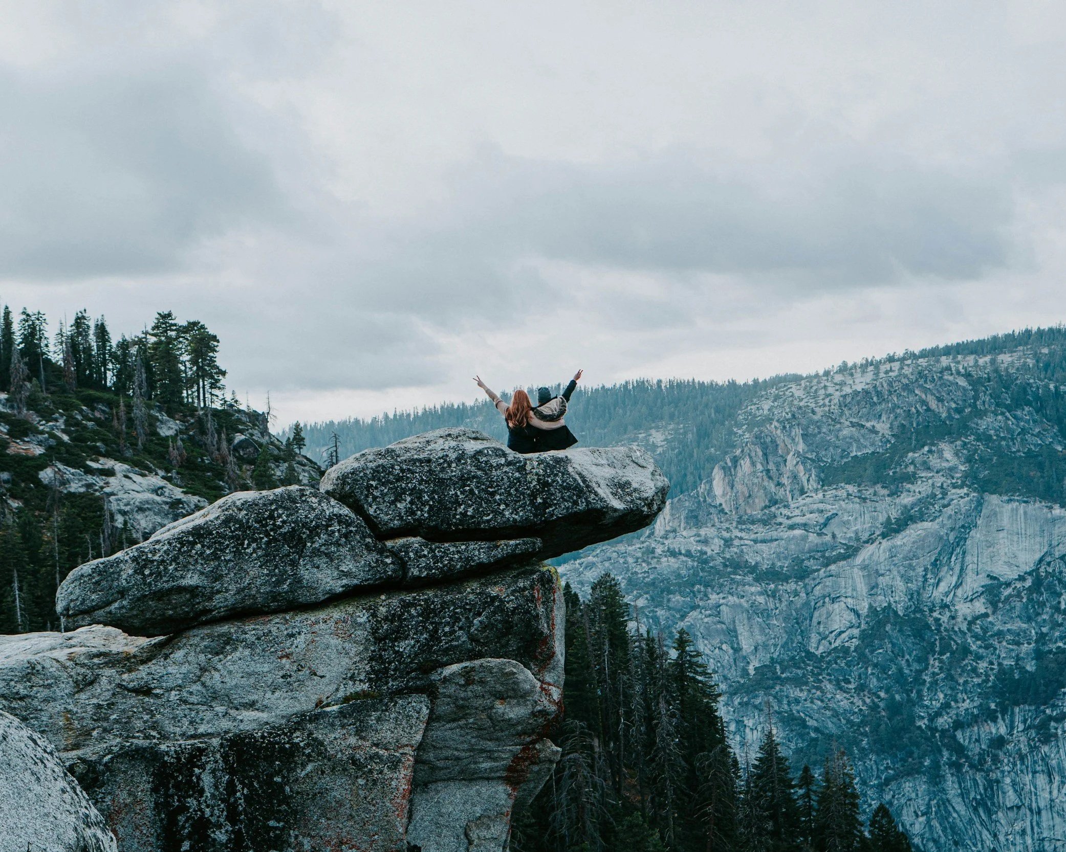 A woman sitting on a large rock formation in a forested mountainous landscape, raising her arms in celebration, under an overcast sky.