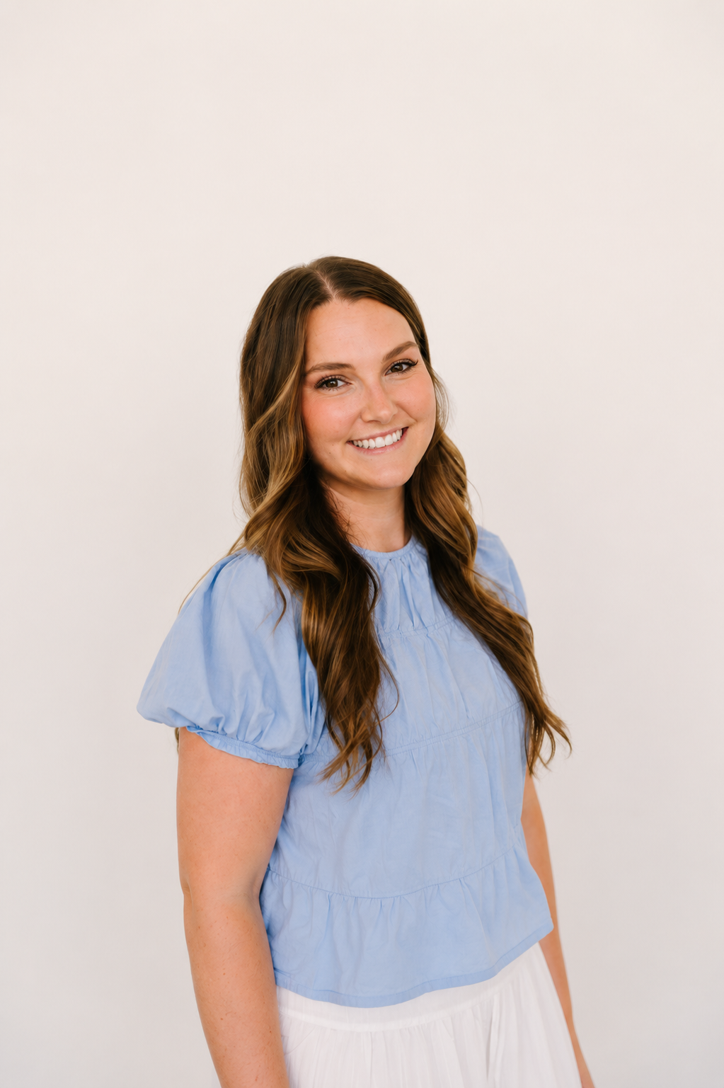 A woman with long brown hair, smiling and posing , wearing a blue short-sleeved top and white pants, standing against a plain white background.