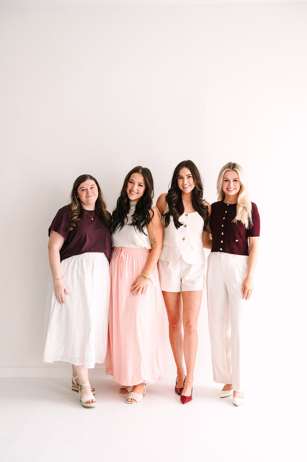 Four women standing together against a plain white background, smiling and dressed in elegant casual attire.