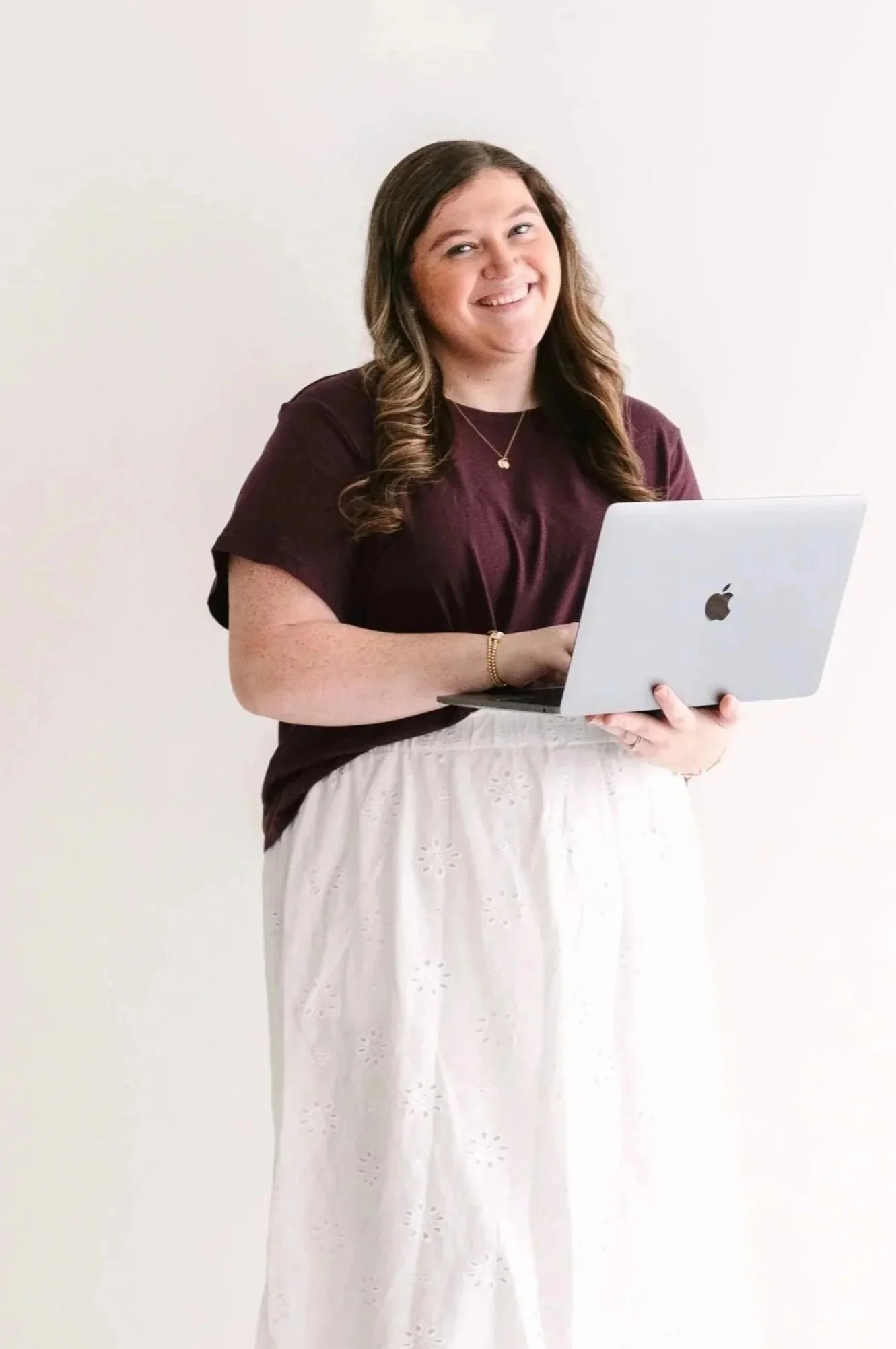 Woman with long brown hair smiling while holding a silver MacBook laptop, standing against a plain white wall.