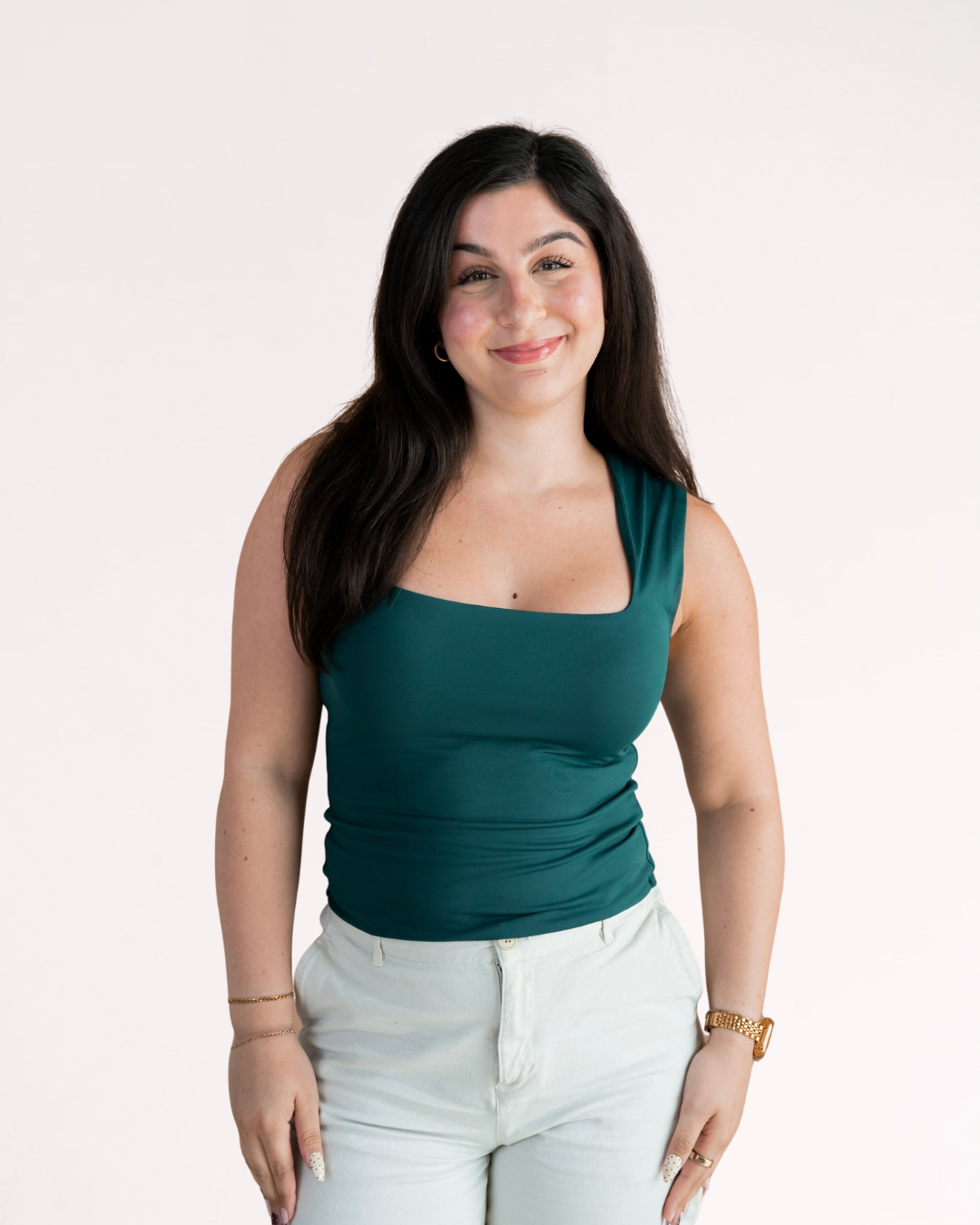 A woman with long brown hair, smiling and posing , wearing a blue short-sleeved top and white pants, standing against a plain white background.