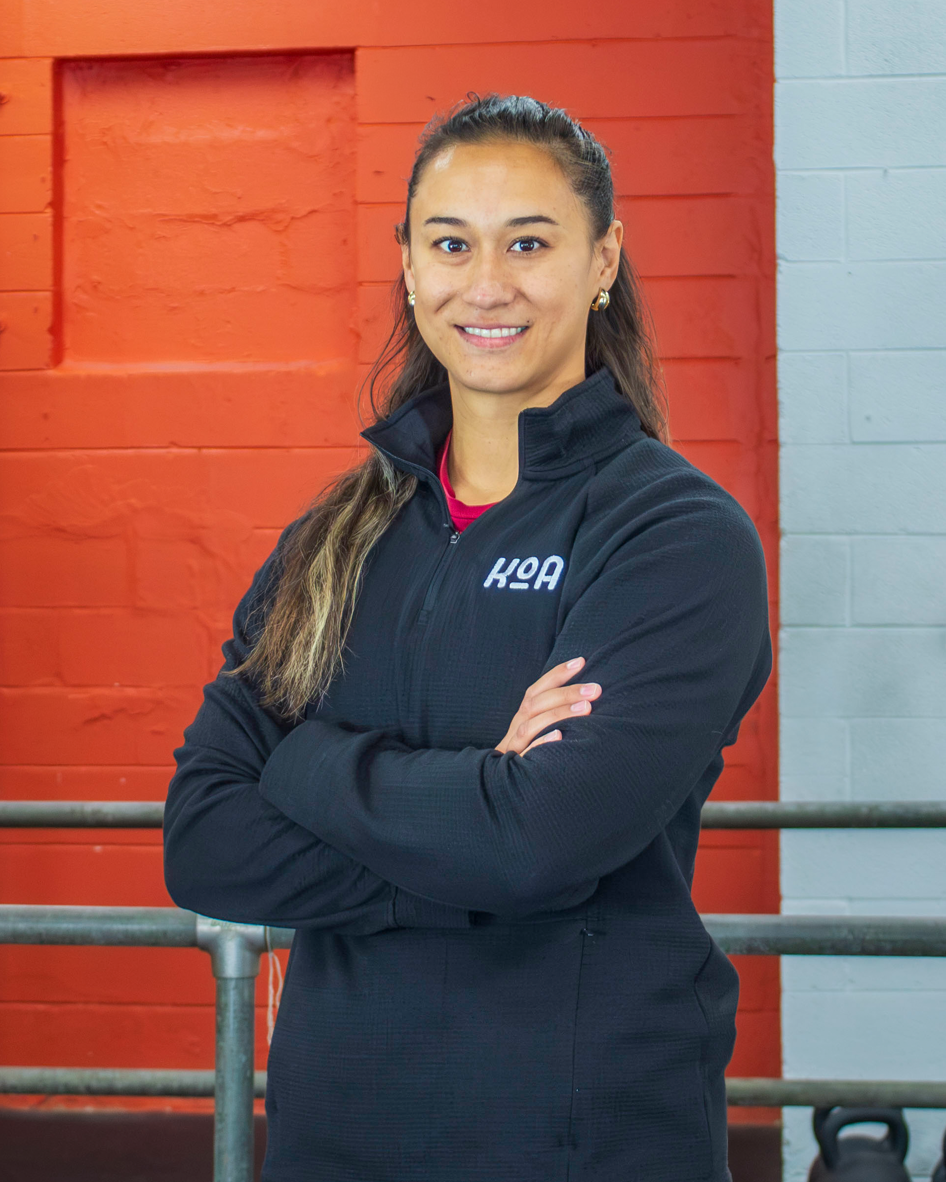A smiling woman standing with arms crossed in a gym with black and yellow weight plates in the background.