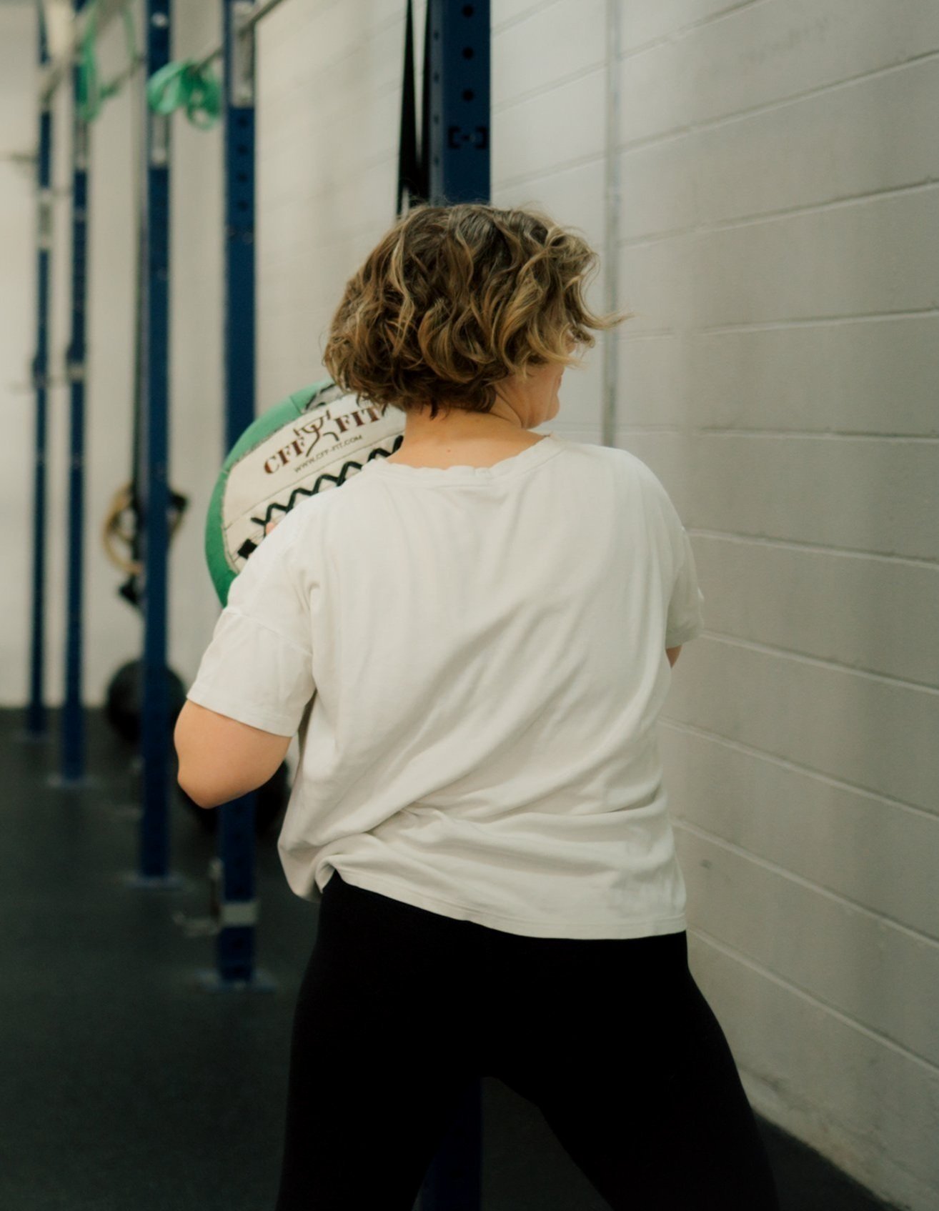 Person with curly hair in a white shirt holding a medicine ball in a gym.