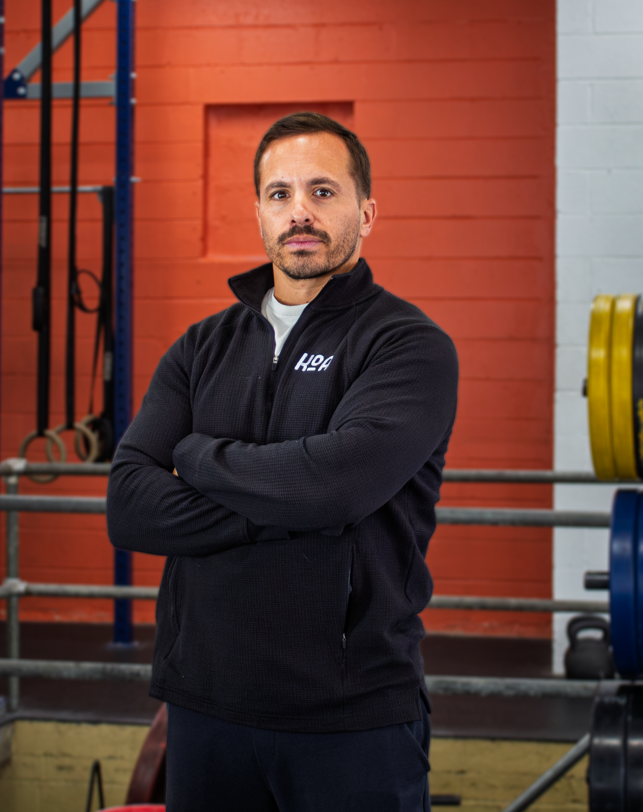 A man with short dark hair and a tattoo on his left arm, smiling with arms crossed, standing in a gym with weightlifting equipment in the background.