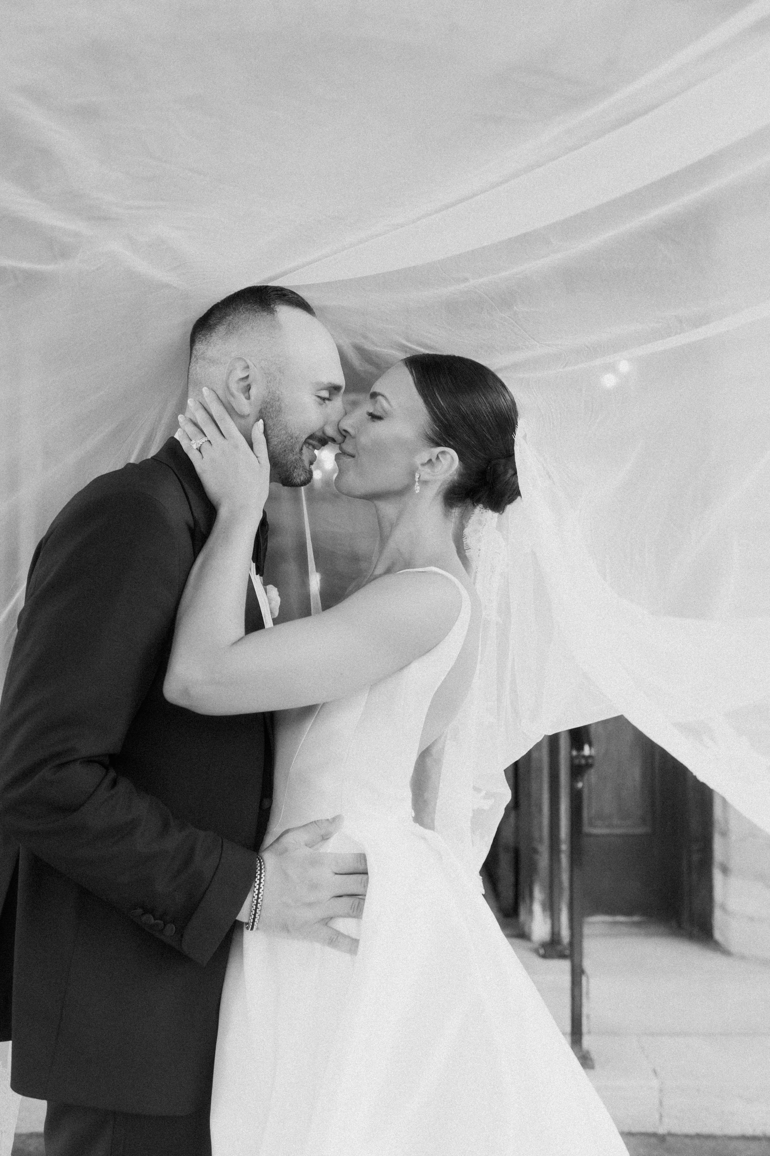 A black and white photograph of a happy couple in wedding attire, sharing an intimate moment under a wedding veil outdoors.