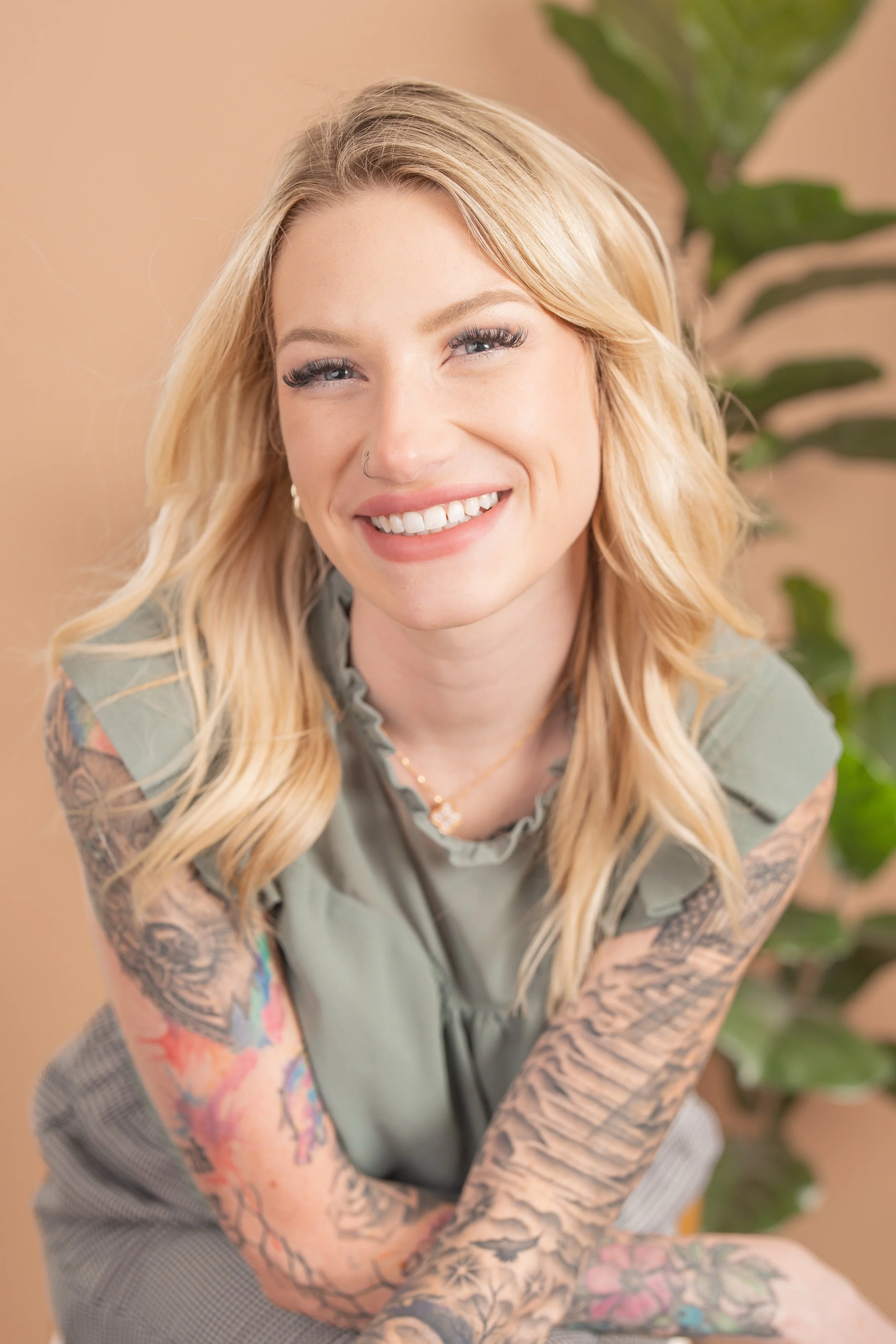 A young woman with blonde hair and tattoos on her arms, smiling and looking at the camera, with a green plant in the background.