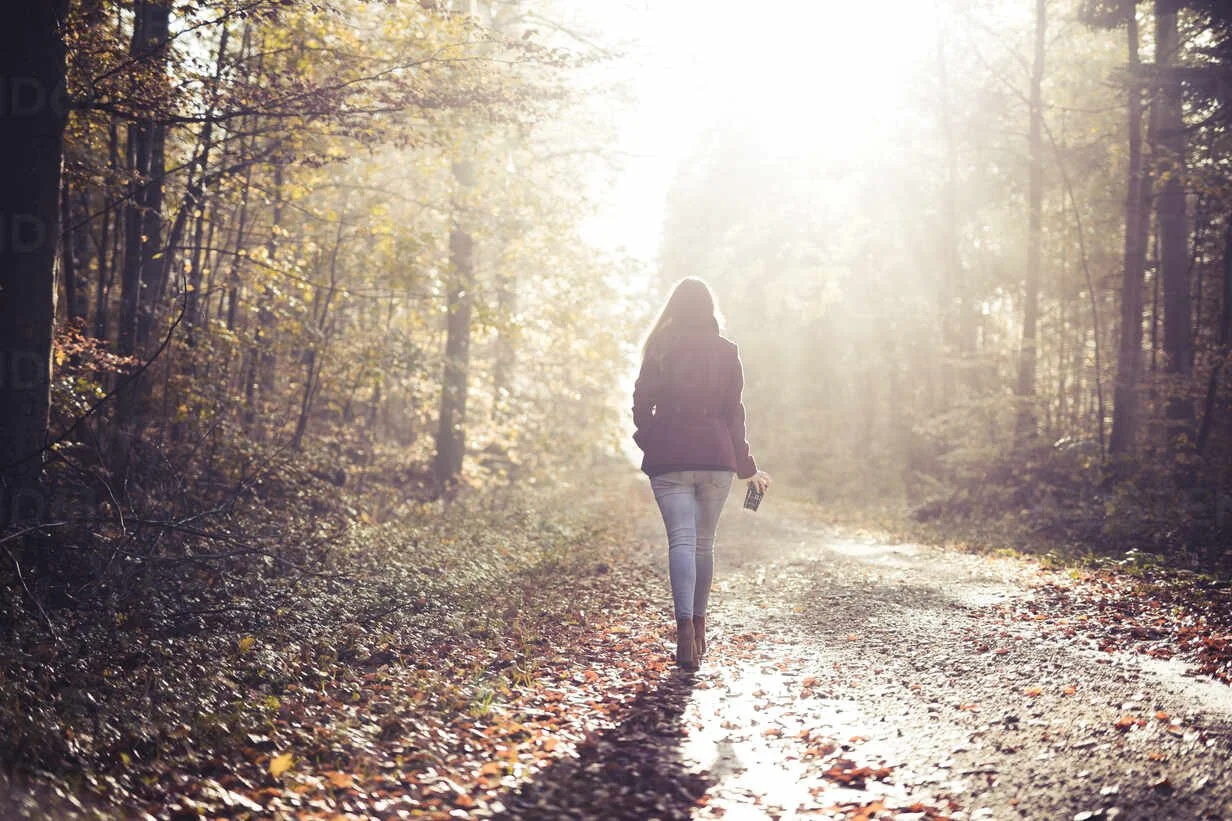 A woman walking down a forest trail surrounded by fall foliage, holding a camera in her right hand, with sunlight shining through the trees.