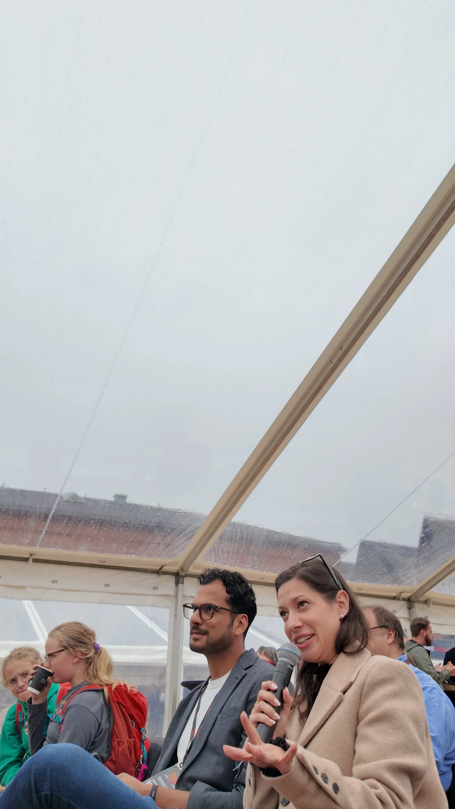 People sitting under a clear tent roof during an event, one woman holding a microphone and talking, with others listening.