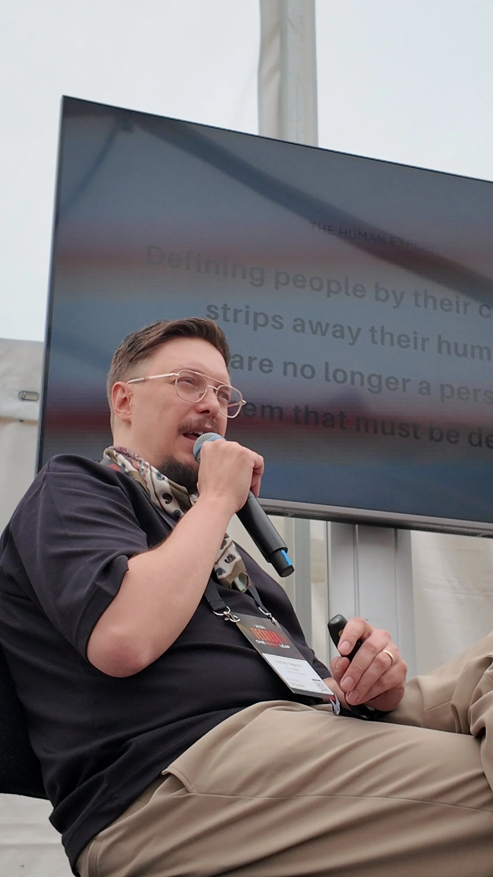 Man holding microphone speaking in front of a screen with text, wearing glasses and a scarf, indoors.