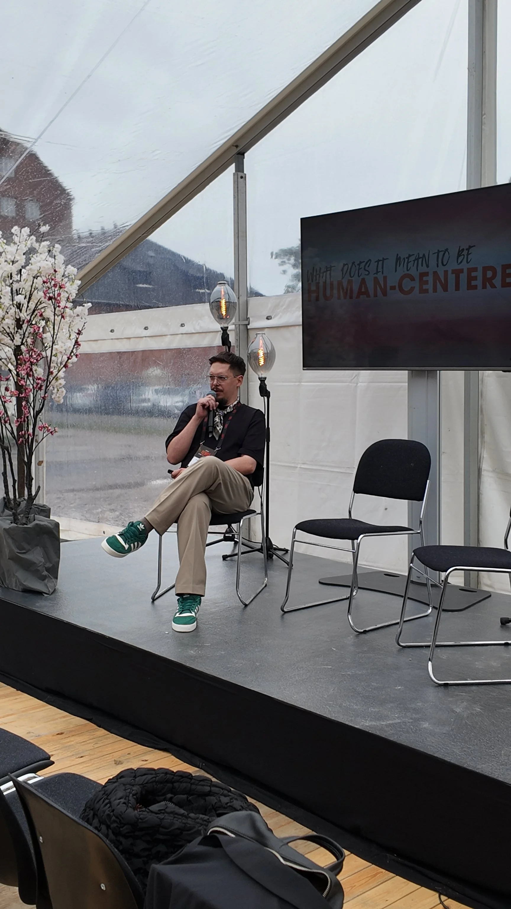 A person sitting on a stage speaking into a microphone, with a large screen displaying the text 'What does it mean to be human-centered?' and a decorative plant nearby.