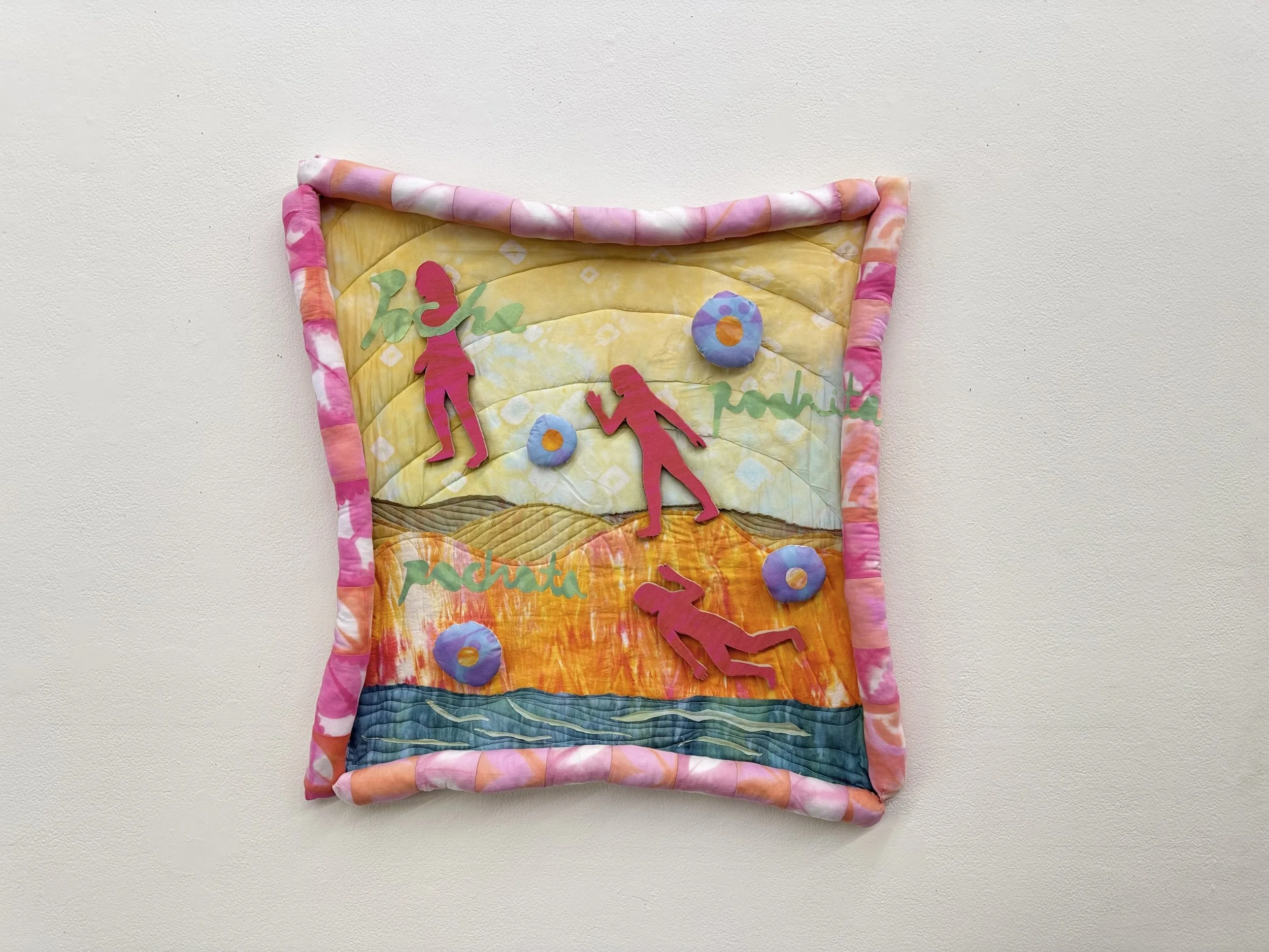 A colorful textile art piece depicting a sunset landscape with silhouettes of two figures, flowers, and the words "hoho pochit" on a white wall.