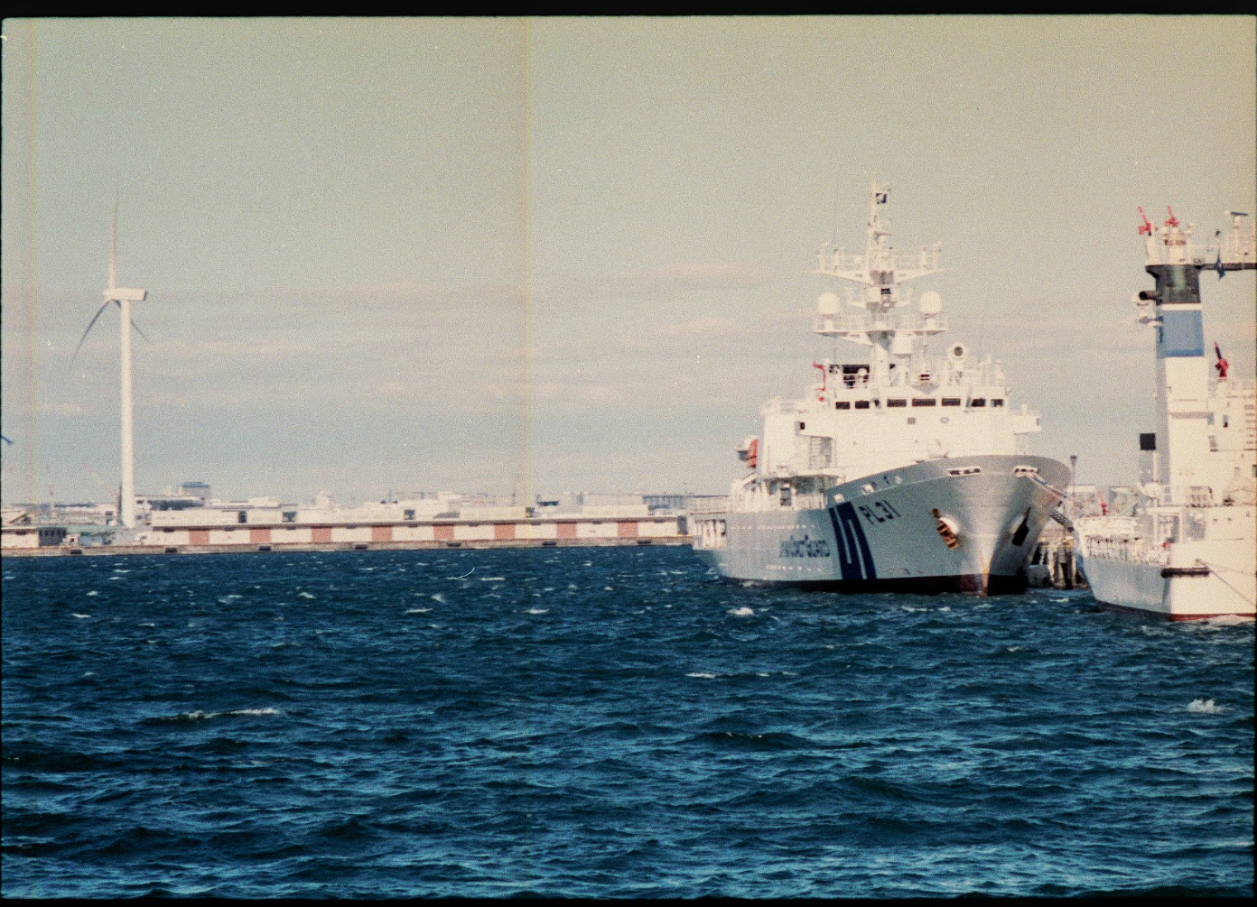 A large white naval ship docked at a port with wind turbines in the background, and another ship partially visible on the right side.