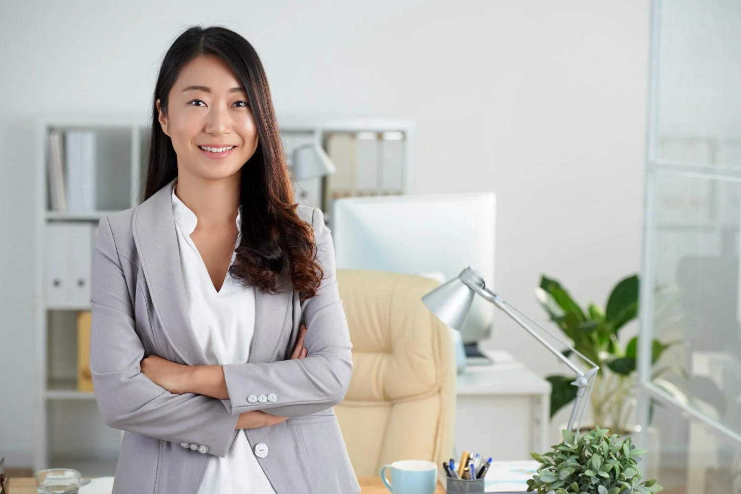 A woman dressed in a light gray blazer and white shirt, smiling with arms crossed, standing in a bright, modern office with a desk, lamp, plants, and bookshelves in the background.